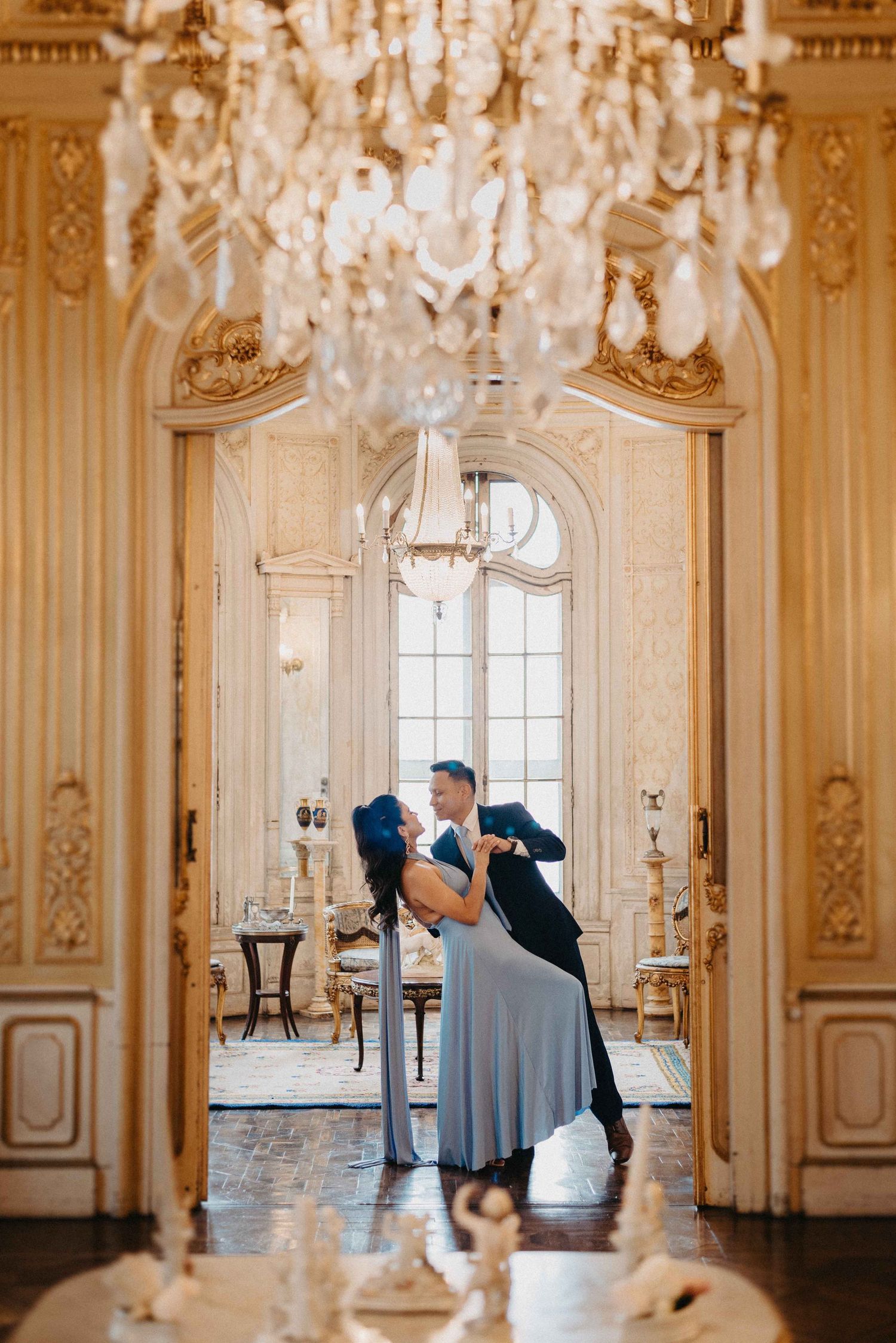 Elegant couple dips in ornate ballroom with crystal chandelier and baroque details.