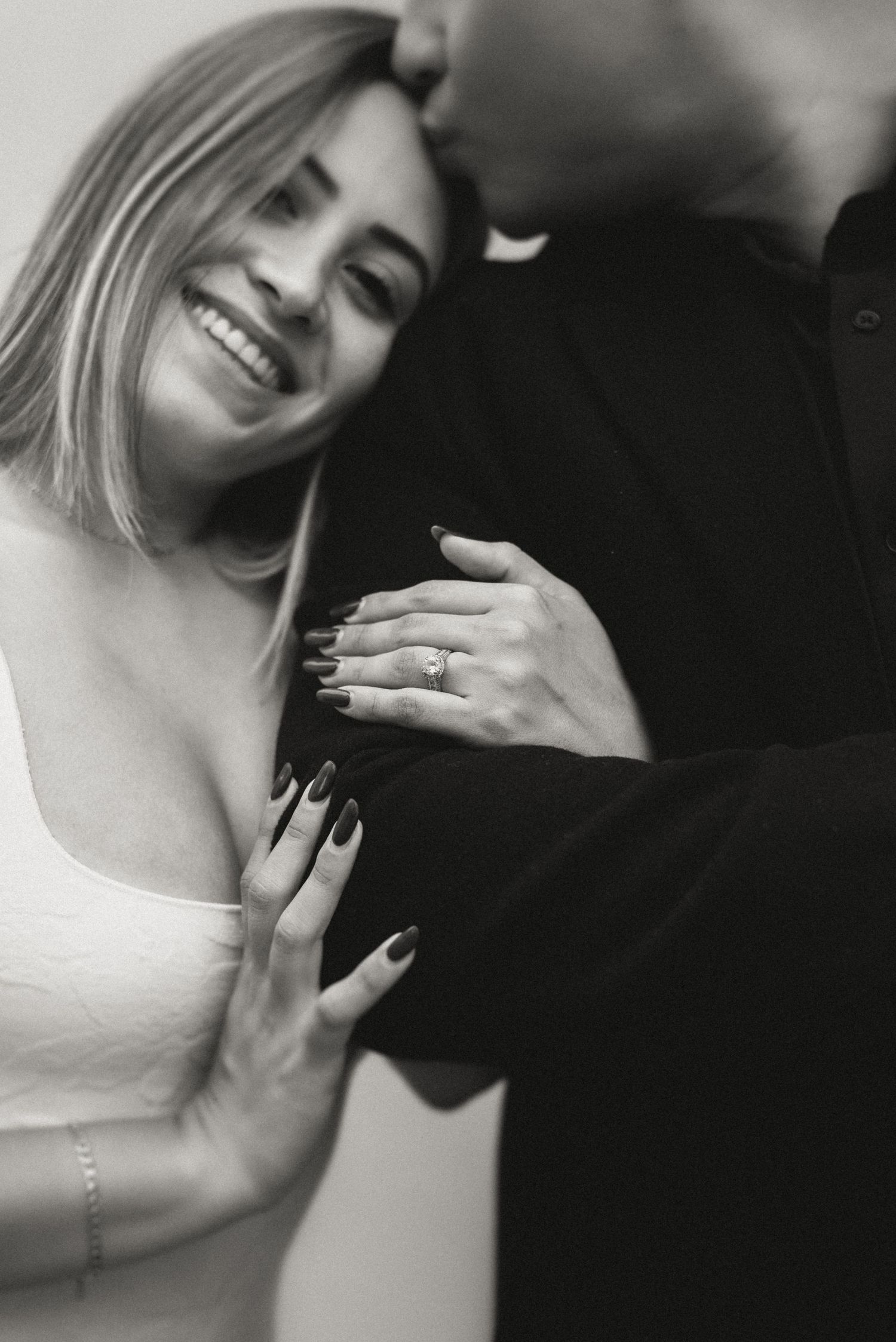 Black and white romantic photo showing a couple embracing with a close-up of their hands displaying an engagement ring.