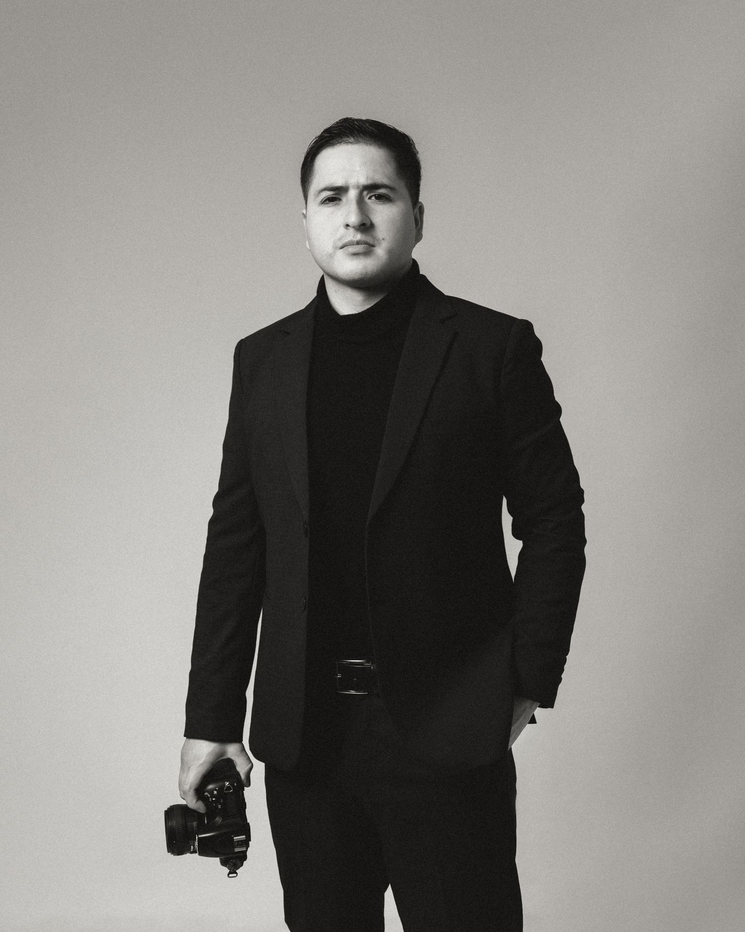 Black and white portrait of a figure in formal attire holding a camera against a neutral background.