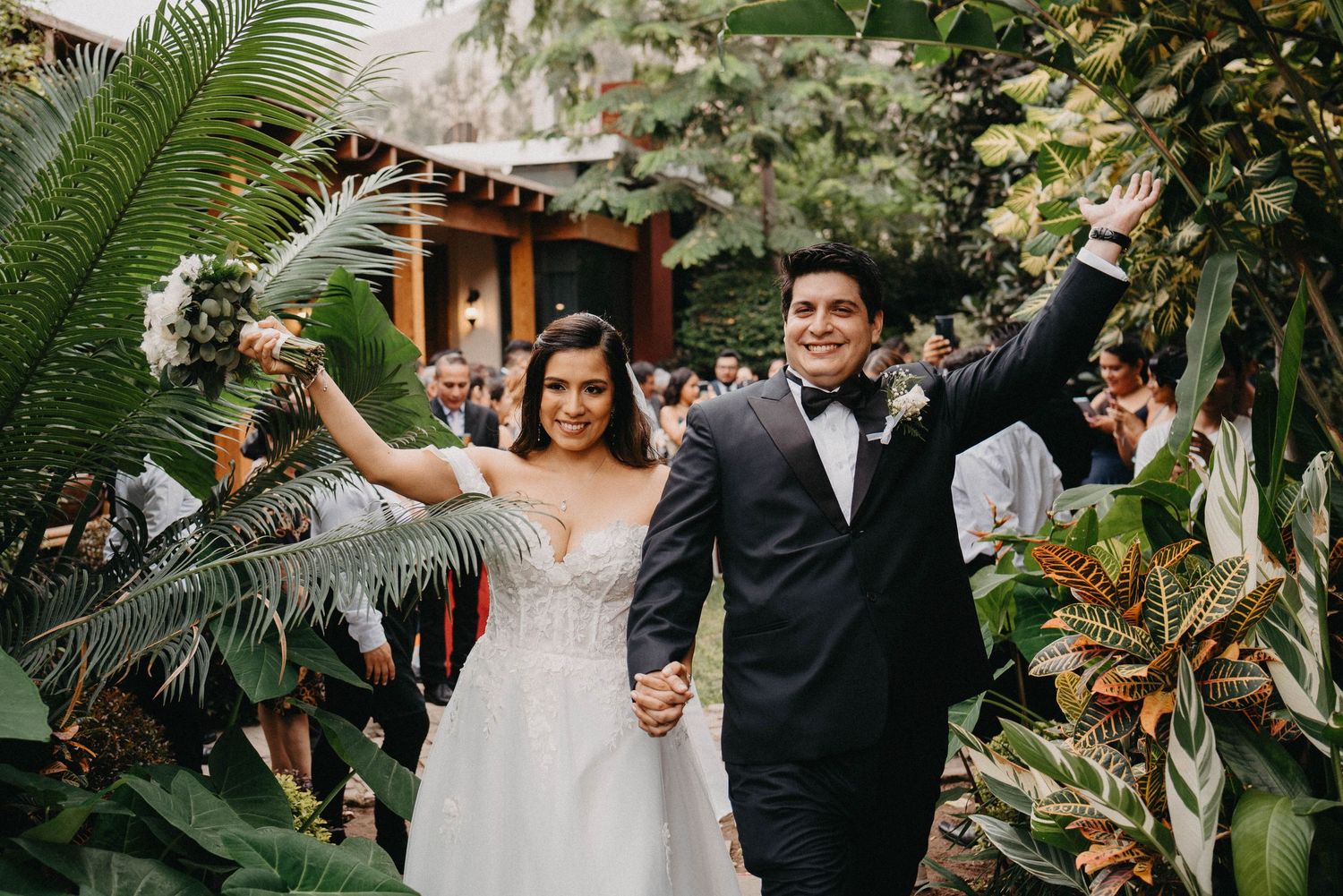 A joyful wedding couple celebrates together in a lush tropical garden setting with palm fronds and greenery.