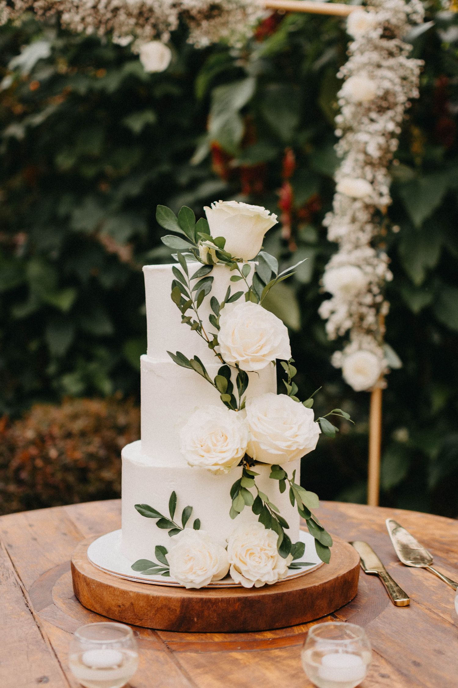 Elegant white wedding cake decorated with fresh white roses and greenery on a rustic wooden table.