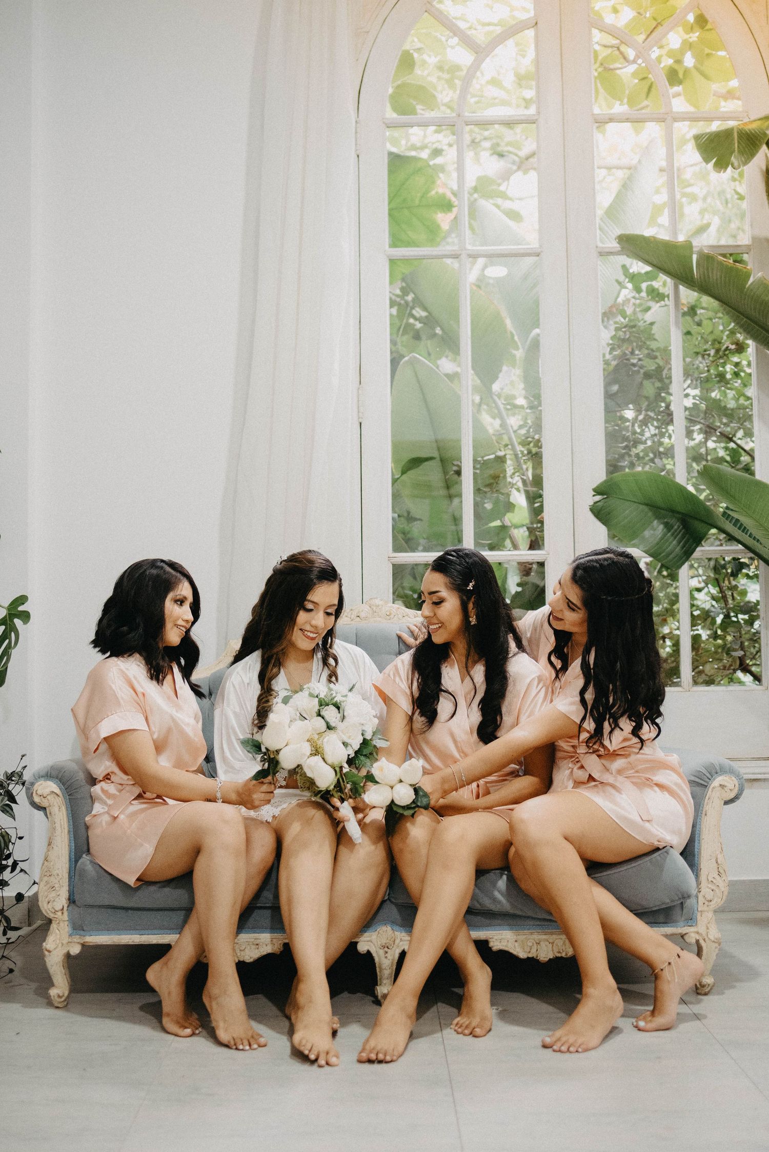 Bridesmaids in pink dresses sit in a row on a white couch by sunny windows holding white floral bouquets.