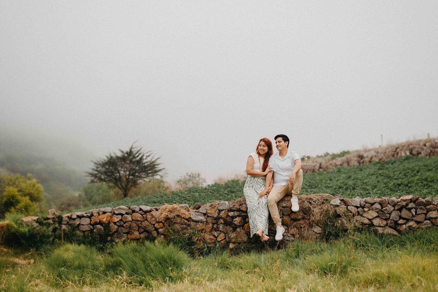 Couple shares romantic moments along stone wall in misty outdoor setting.
