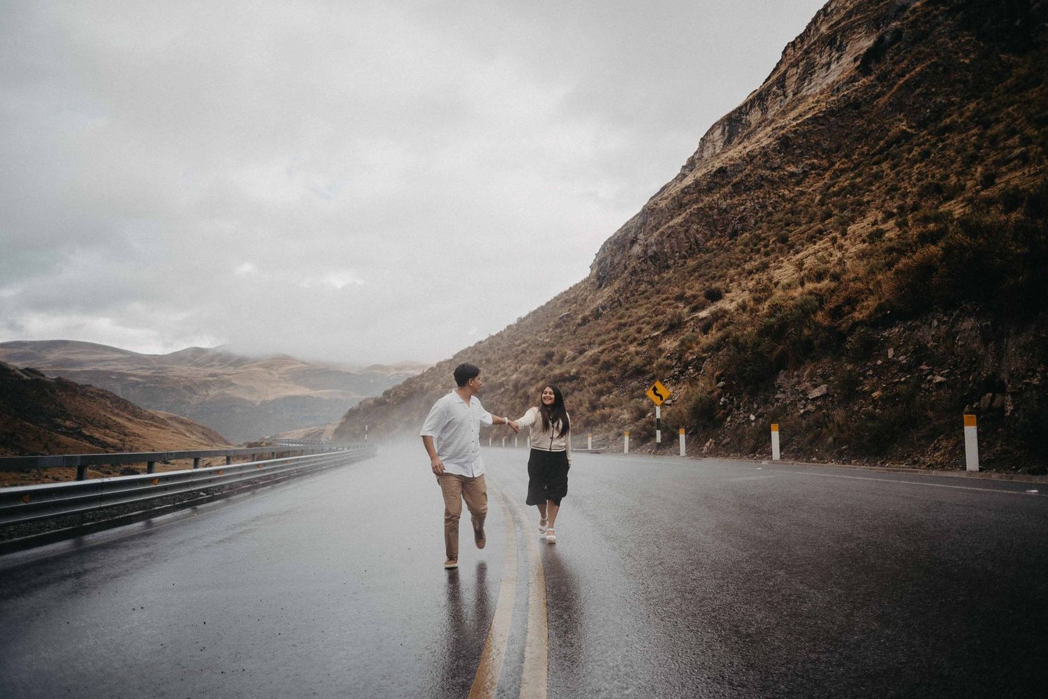 Two people share a joyful moment on a wet mountain road surrounded by misty hills.
