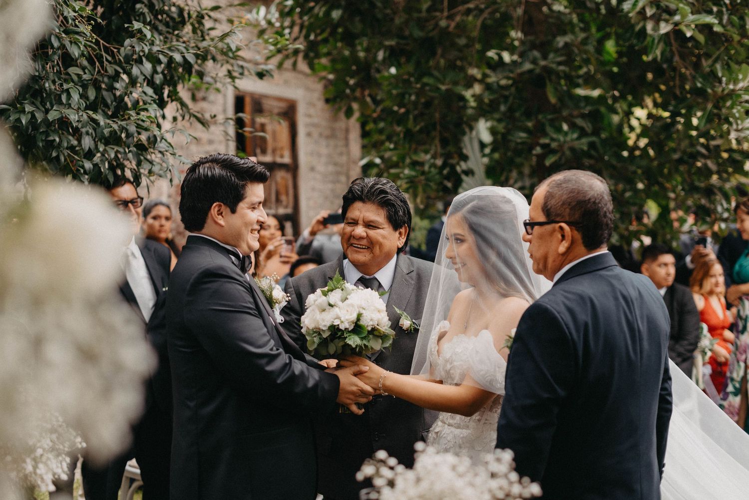 Wedding ceremony guests seated outdoors among trees and greenery.
