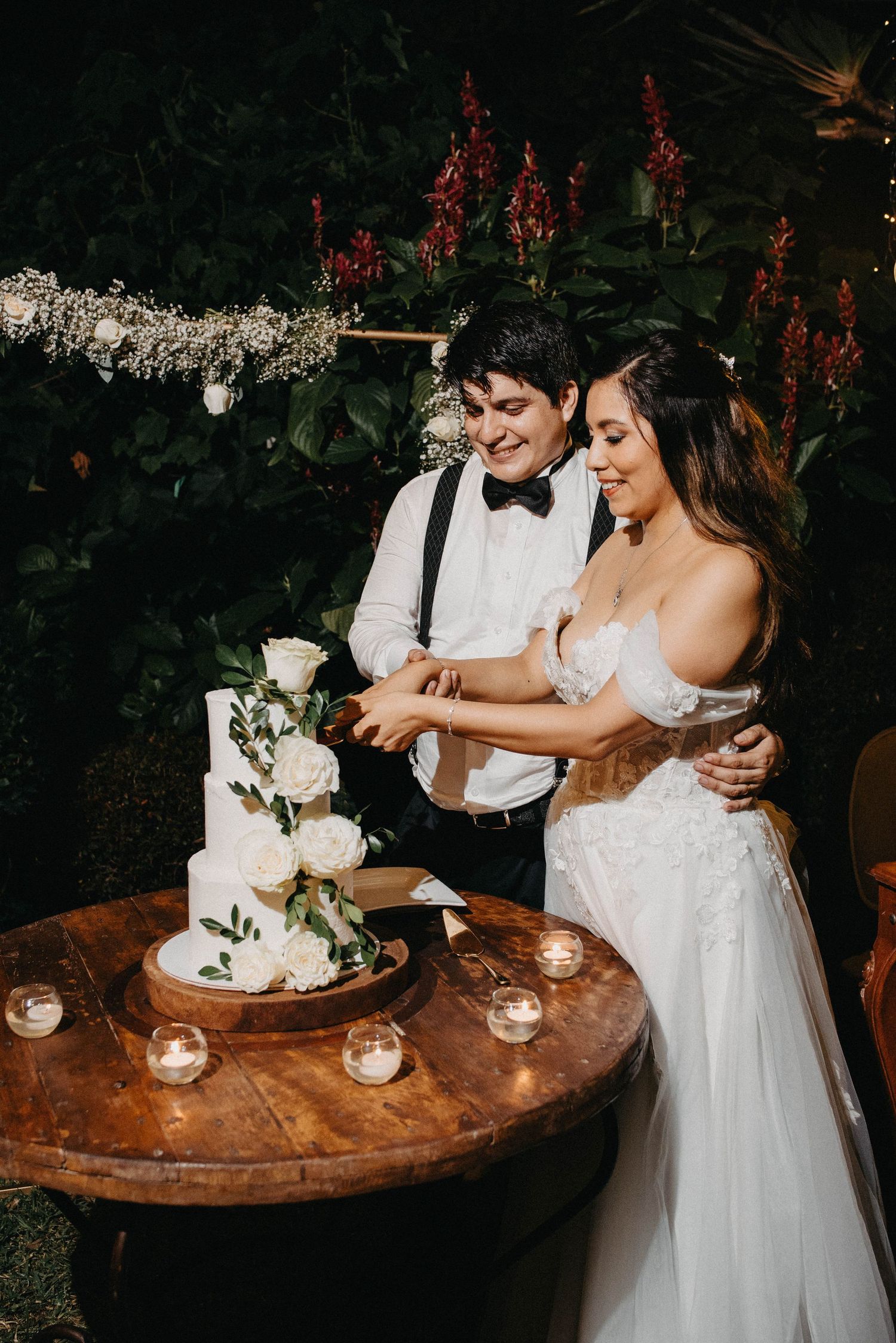 Couple sharing intimate moment while cutting their elegant white wedding cake on wooden table.