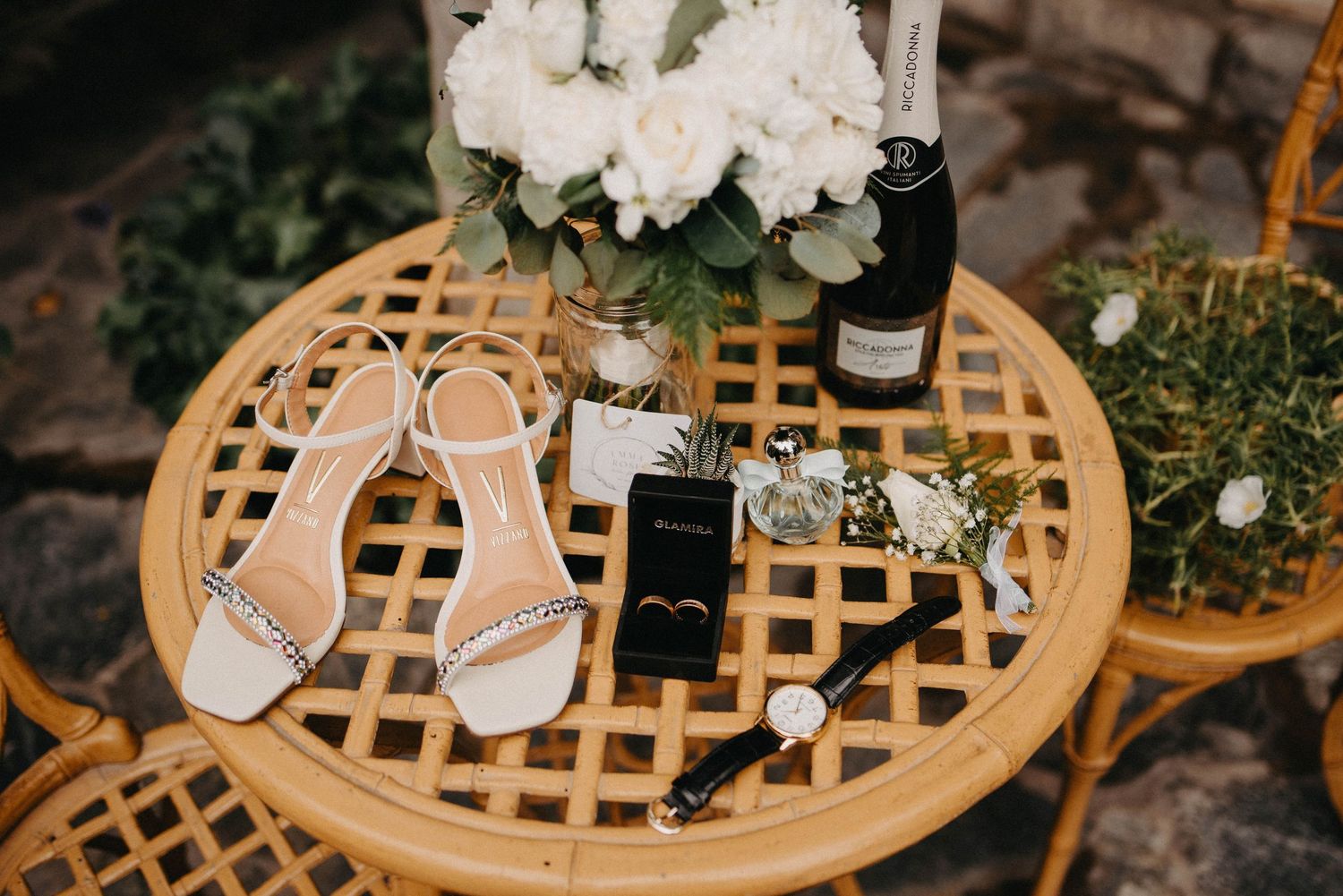 Wedding accessories including white sandals, phone, and jewelry arranged on a round lattice wooden table with flowers.
