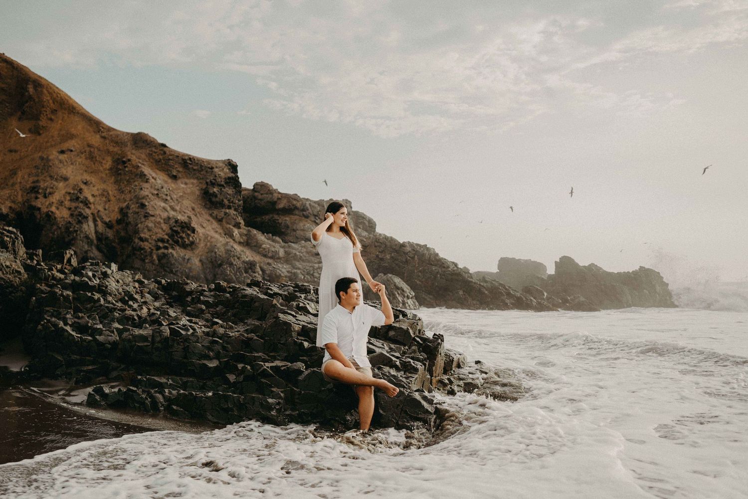Two people playing joyfully on a rocky beach with waves and cliffs in the background.