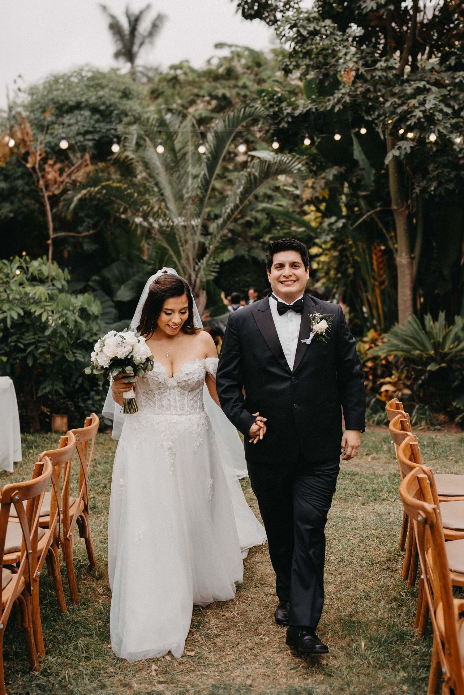 Bride and groom walking down aisle at tropical garden wedding with string lights.