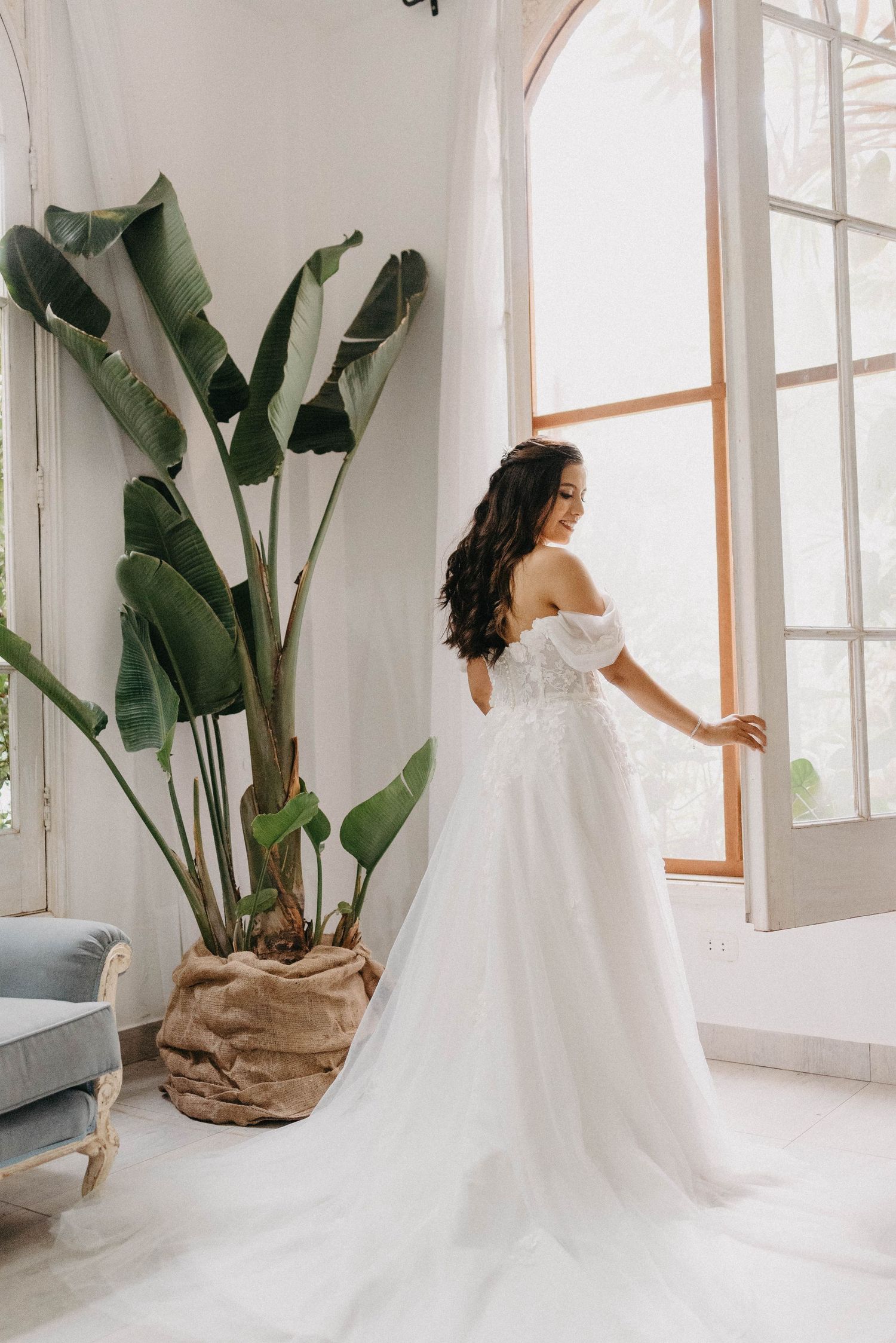 Bride in flowing white wedding gown twirls gracefully in bright sunlit room with large windows.