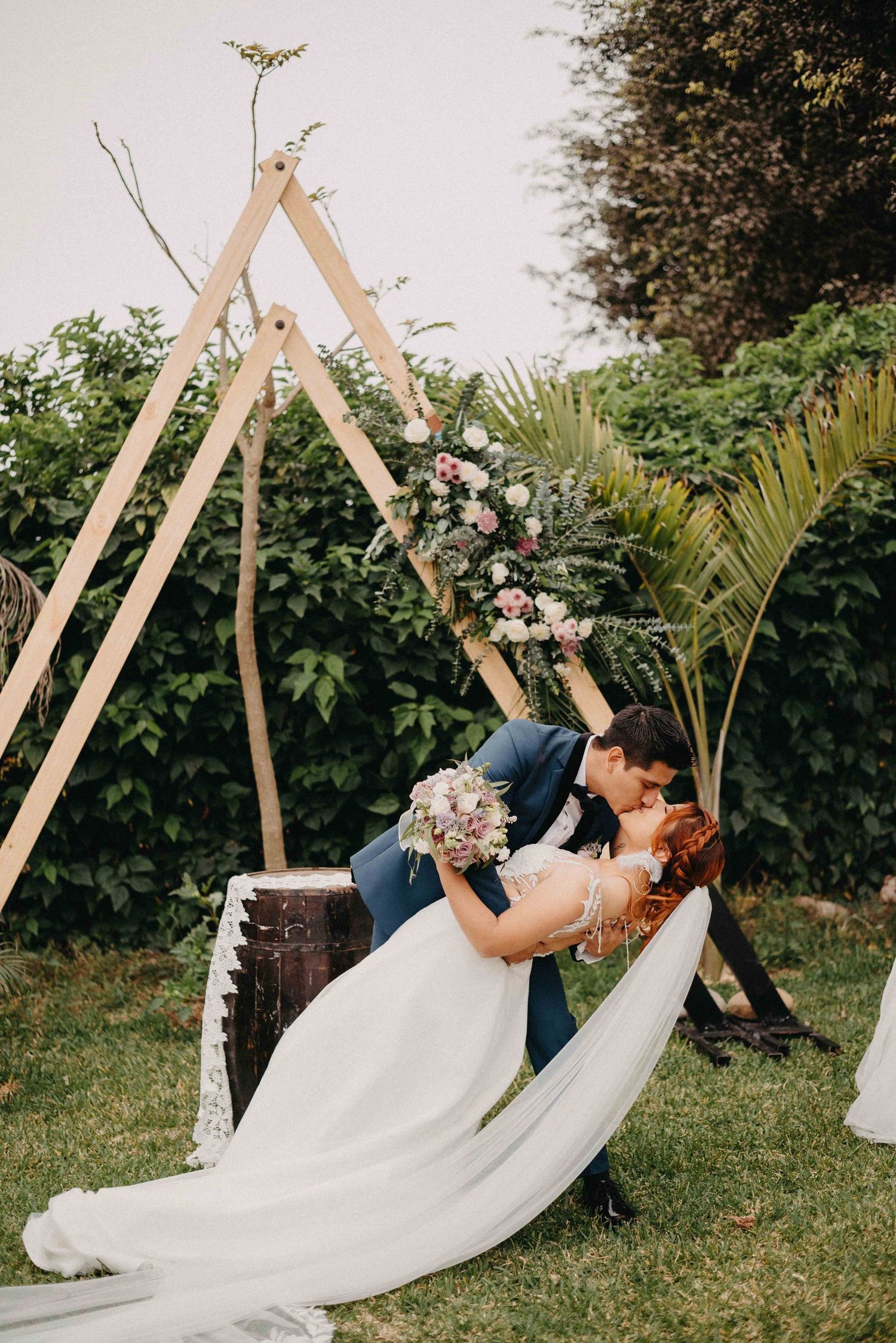 Wedding couple shares romantic dip kiss under wooden triangle arch decorated with flowers in garden setting.