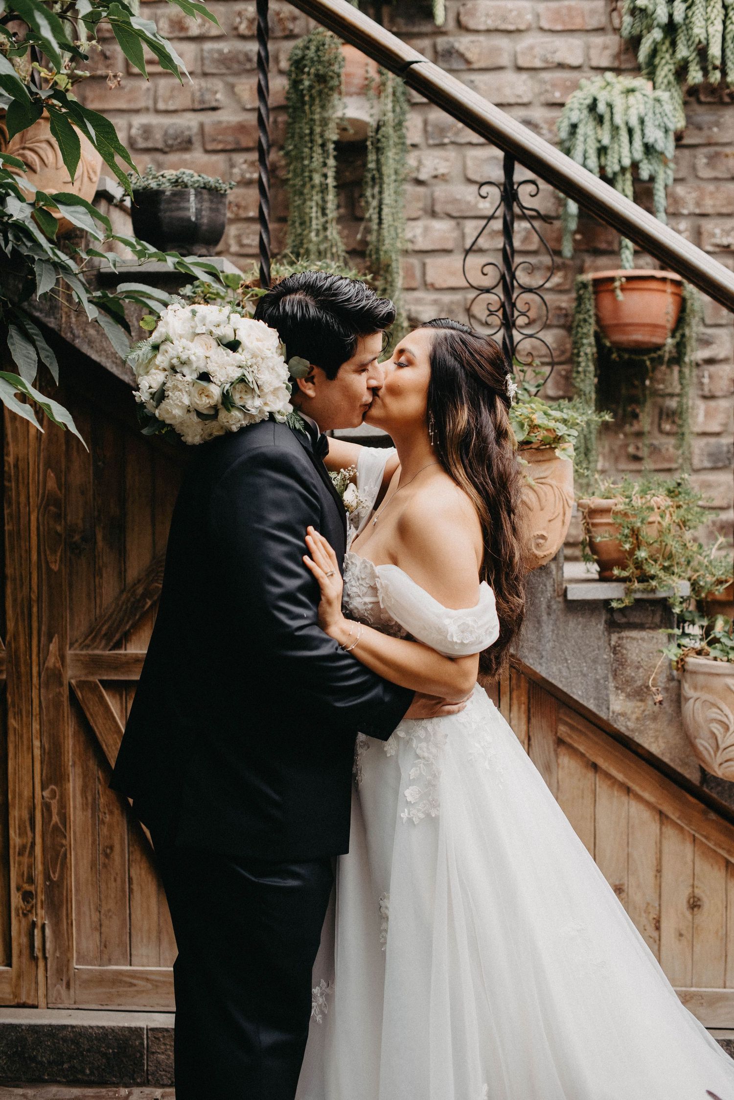 Bride in flowing white dress and groom in black tuxedo share romantic kisses against brick wall backdrop.