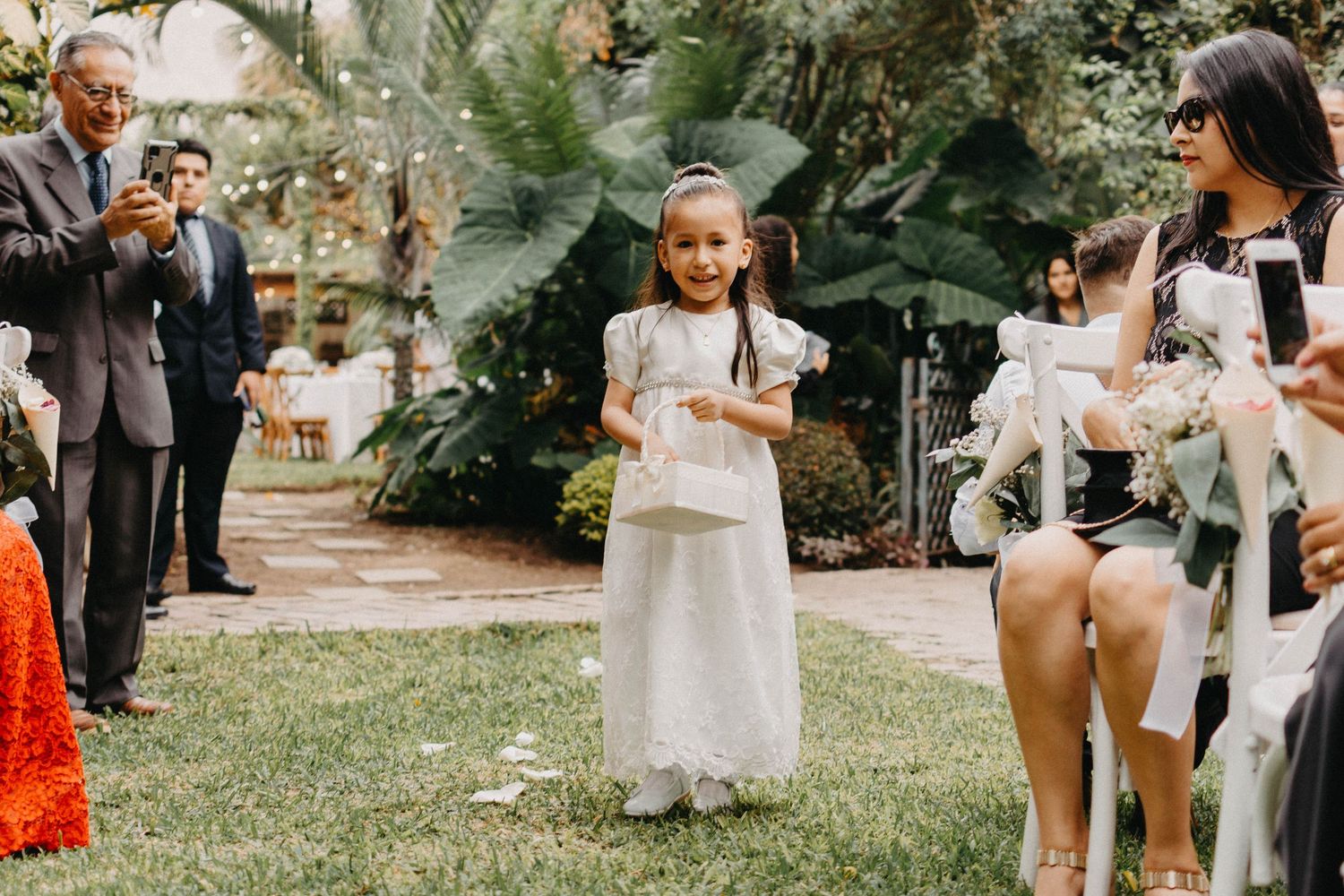 A small child in white dress stands on grass during an outdoor wedding ceremony.