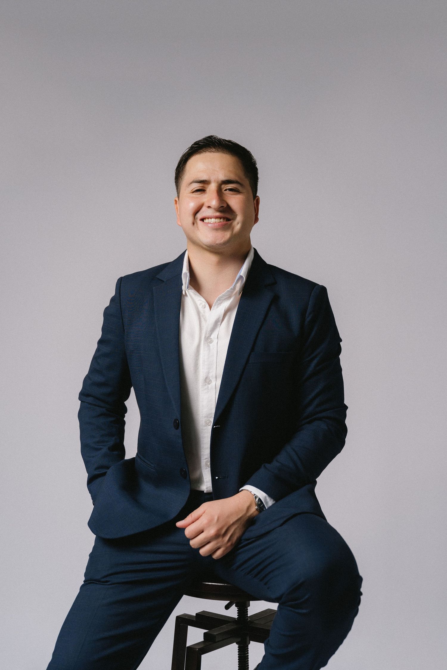 Professional business portrait of person in navy suit sitting on stool against white background with warm smile.