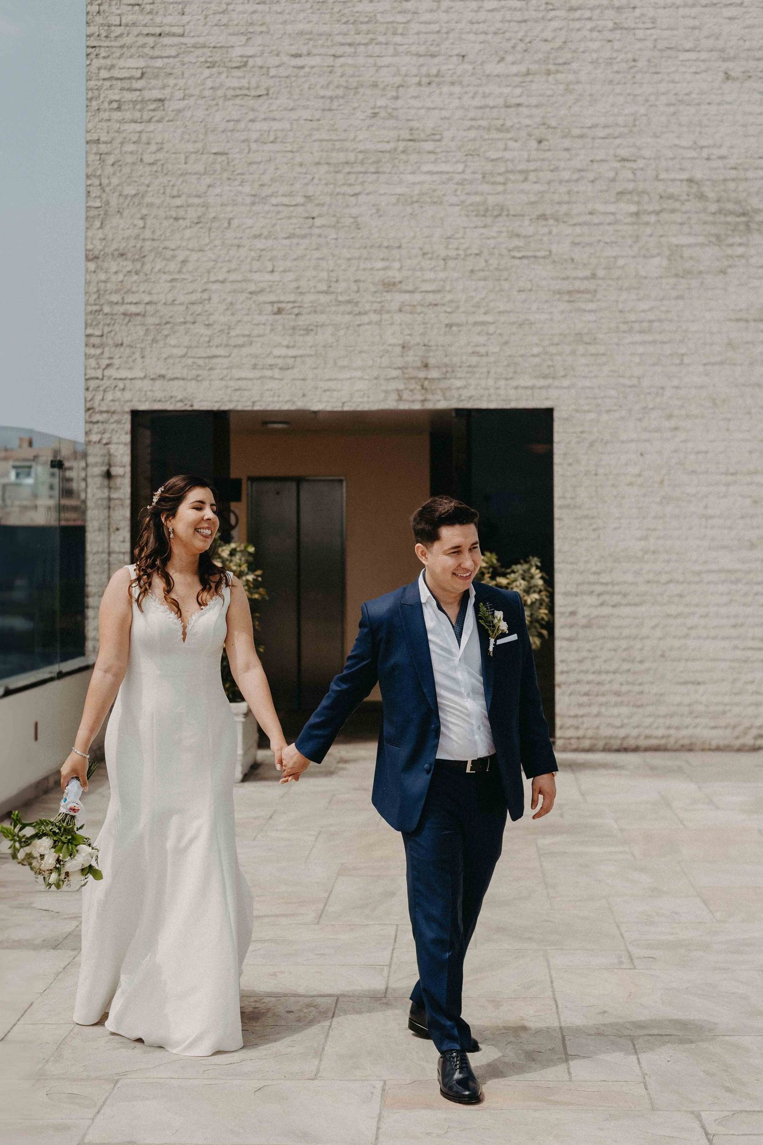 Newlyweds hold hands while walking together outside a modern brick building.
