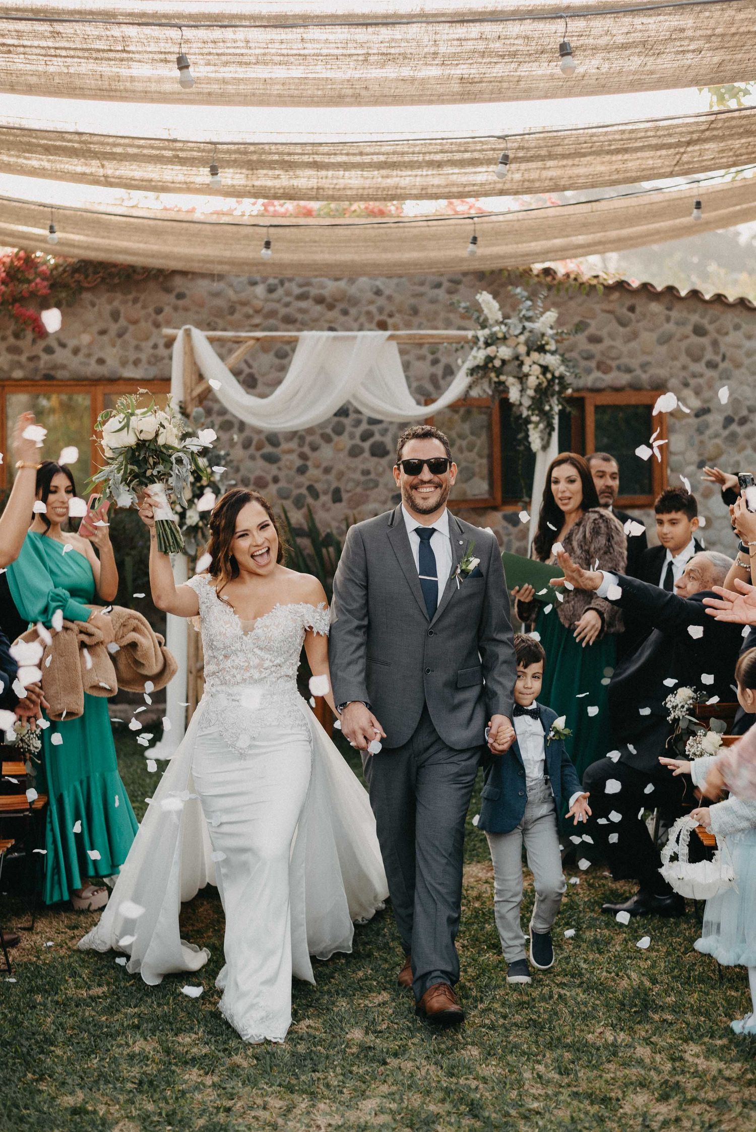 Wedding celebration with guests tossing flower petals at newlyweds under a draped outdoor tent.