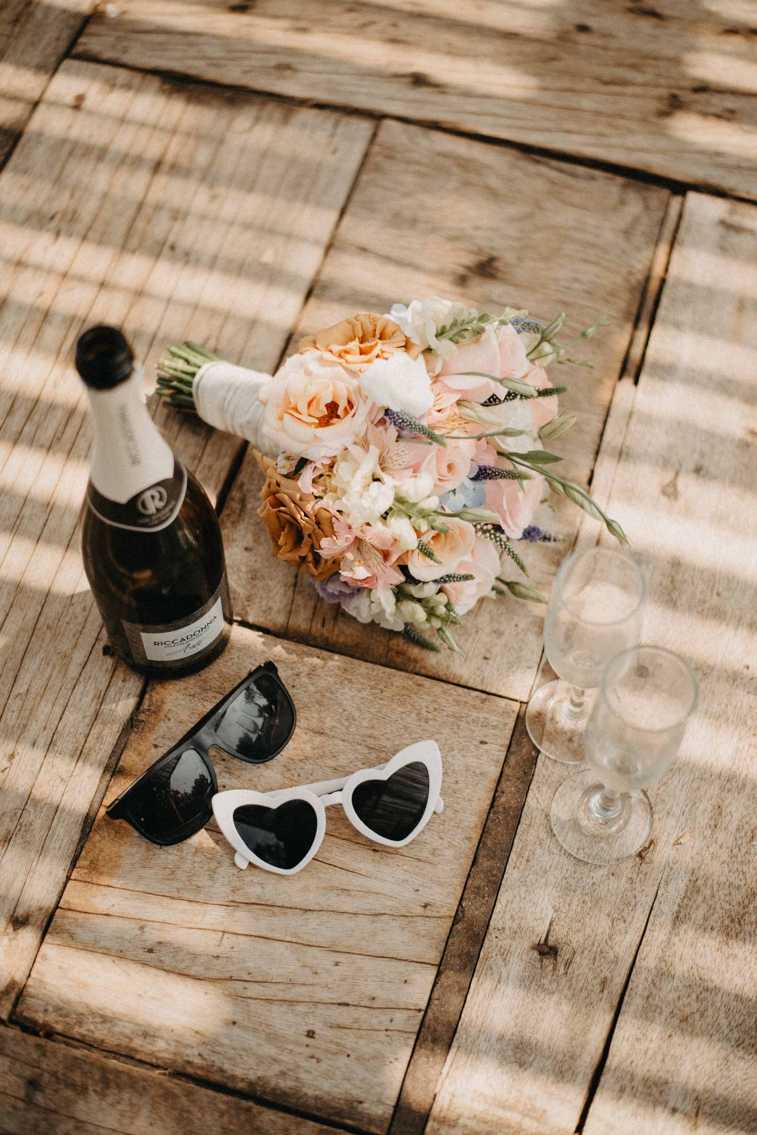 Wedding details with champagne bottle, heart-shaped sunglasses and peach floral bouquet on rustic wooden surface.