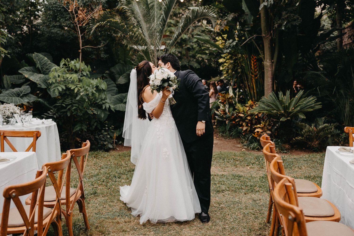 Outdoor wedding ceremony setup with wooden cross-back chairs and lush greenery.
