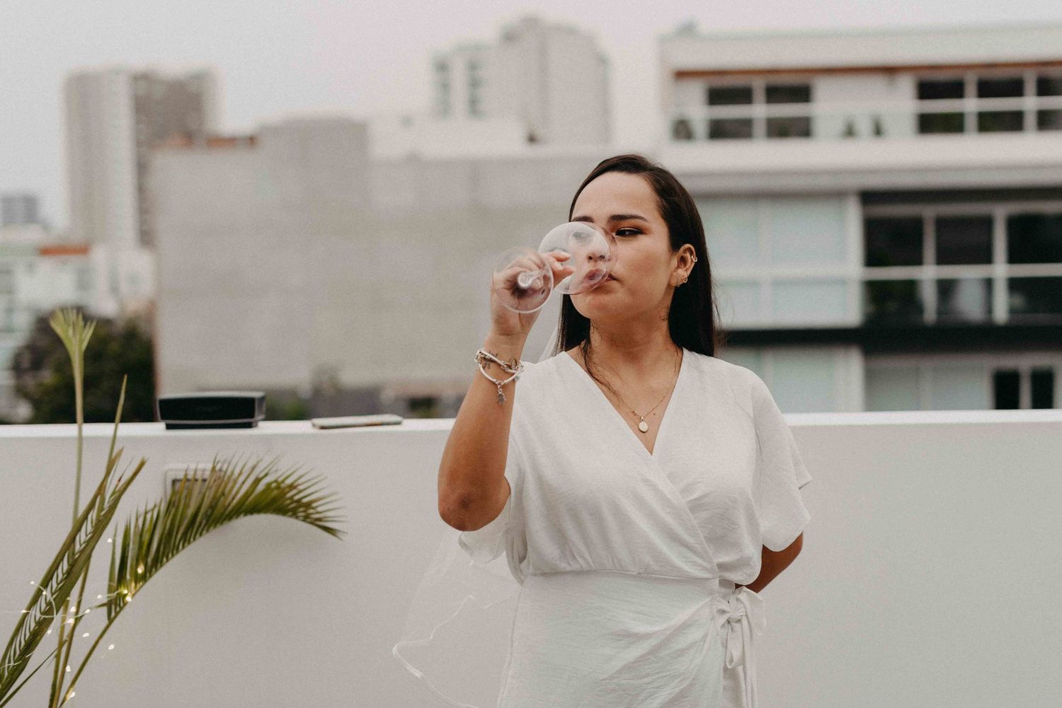 A person in white dress enjoying a drink on an urban rooftop with palm plants nearby.