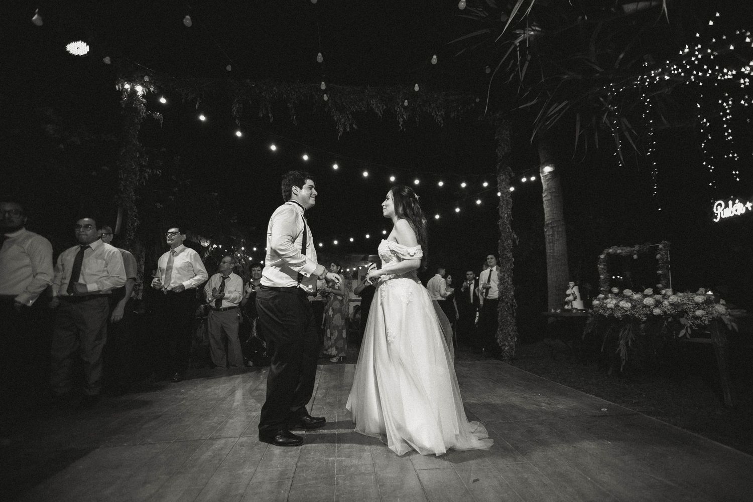 A black and white photo captures a couple sharing their first dance under string lights at their wedding reception.