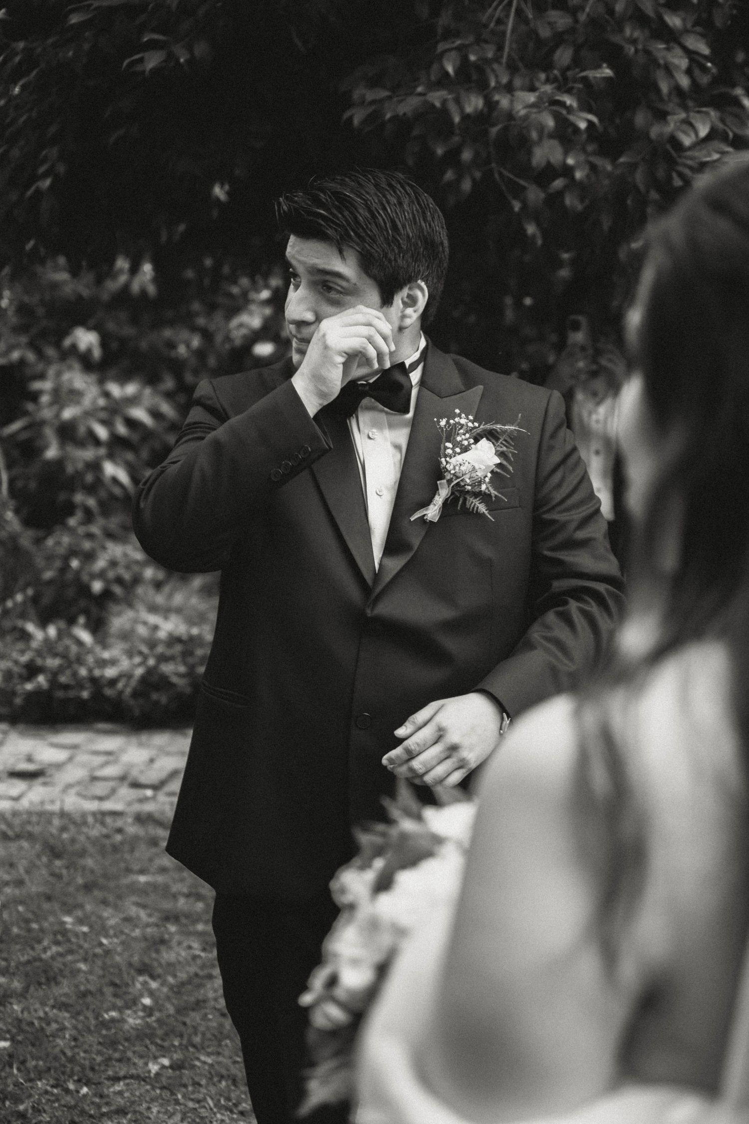 Black and white photo of emotional groom wiping tears during wedding ceremony.