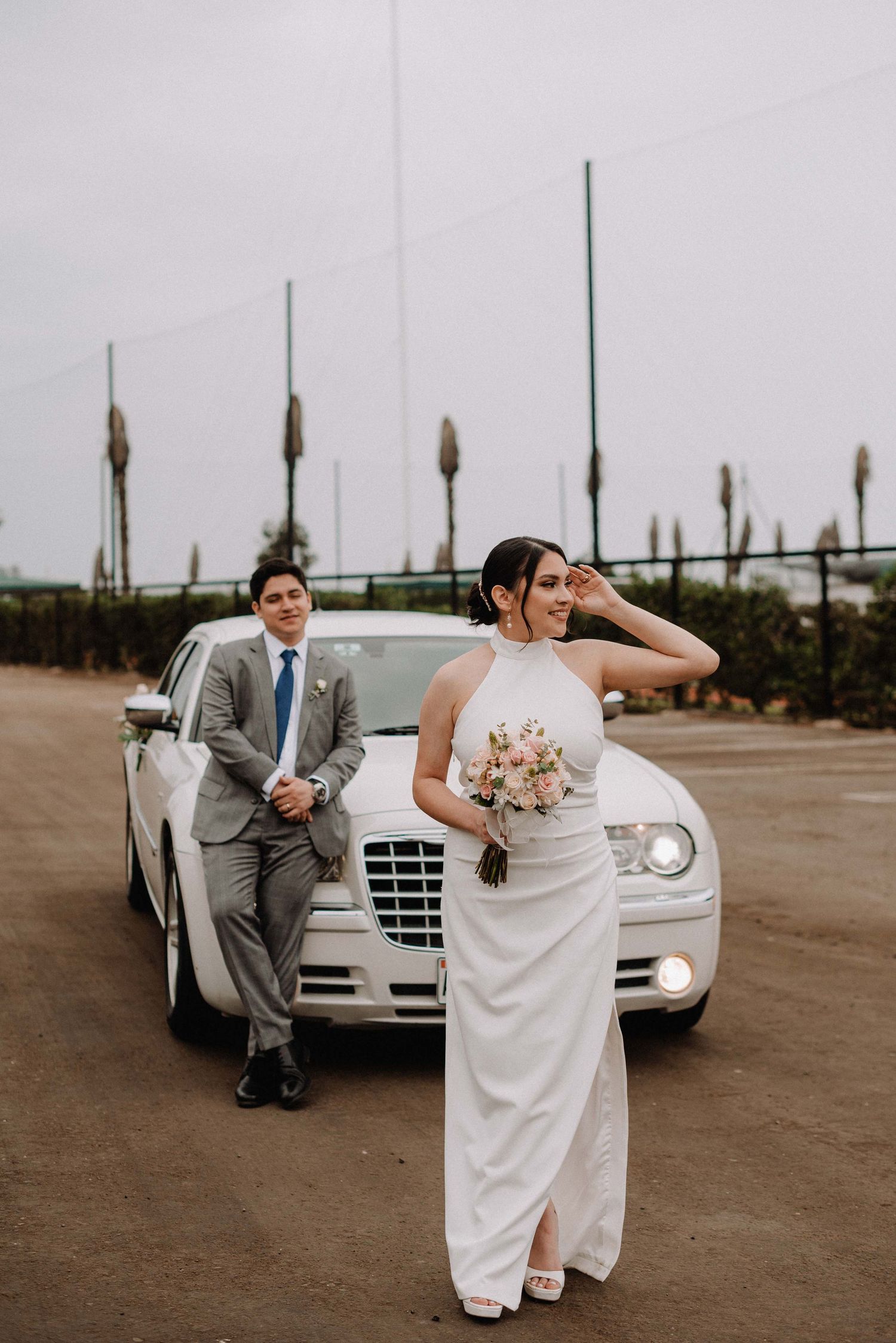 A couple poses with white car featuring the bride in elegant white dress holding bouquet and groom in gray suit.