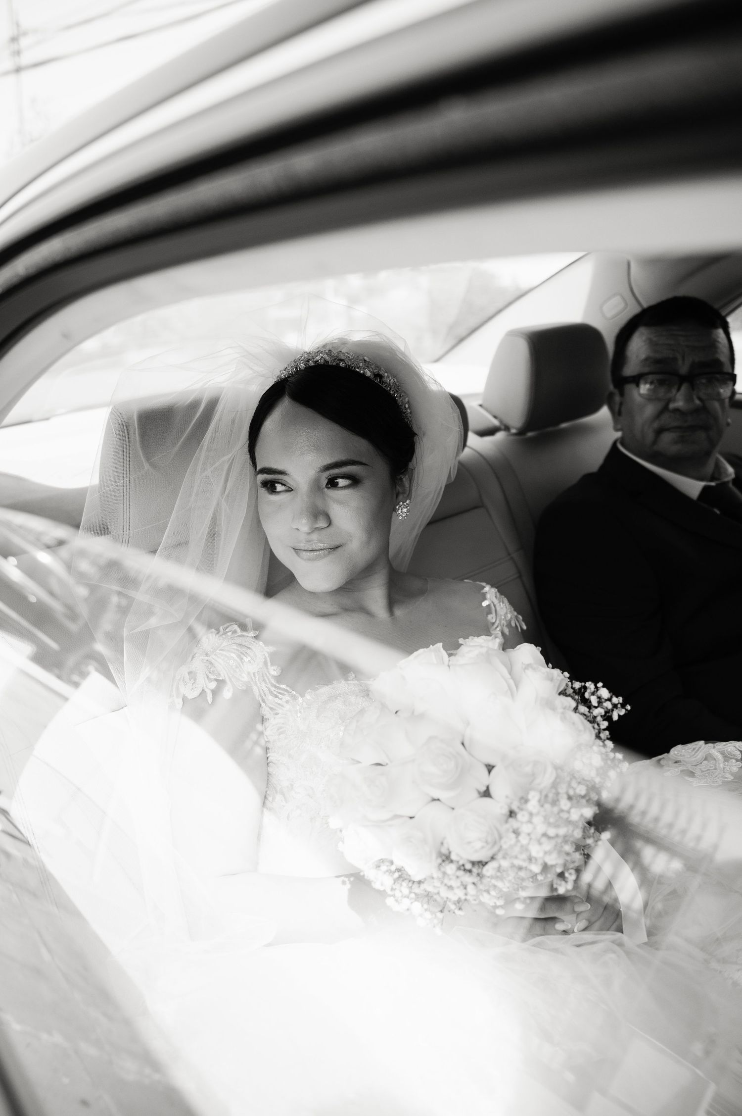 A bride wearing a white veil and dress sits in a car on her wedding day, captured in black and white photography.