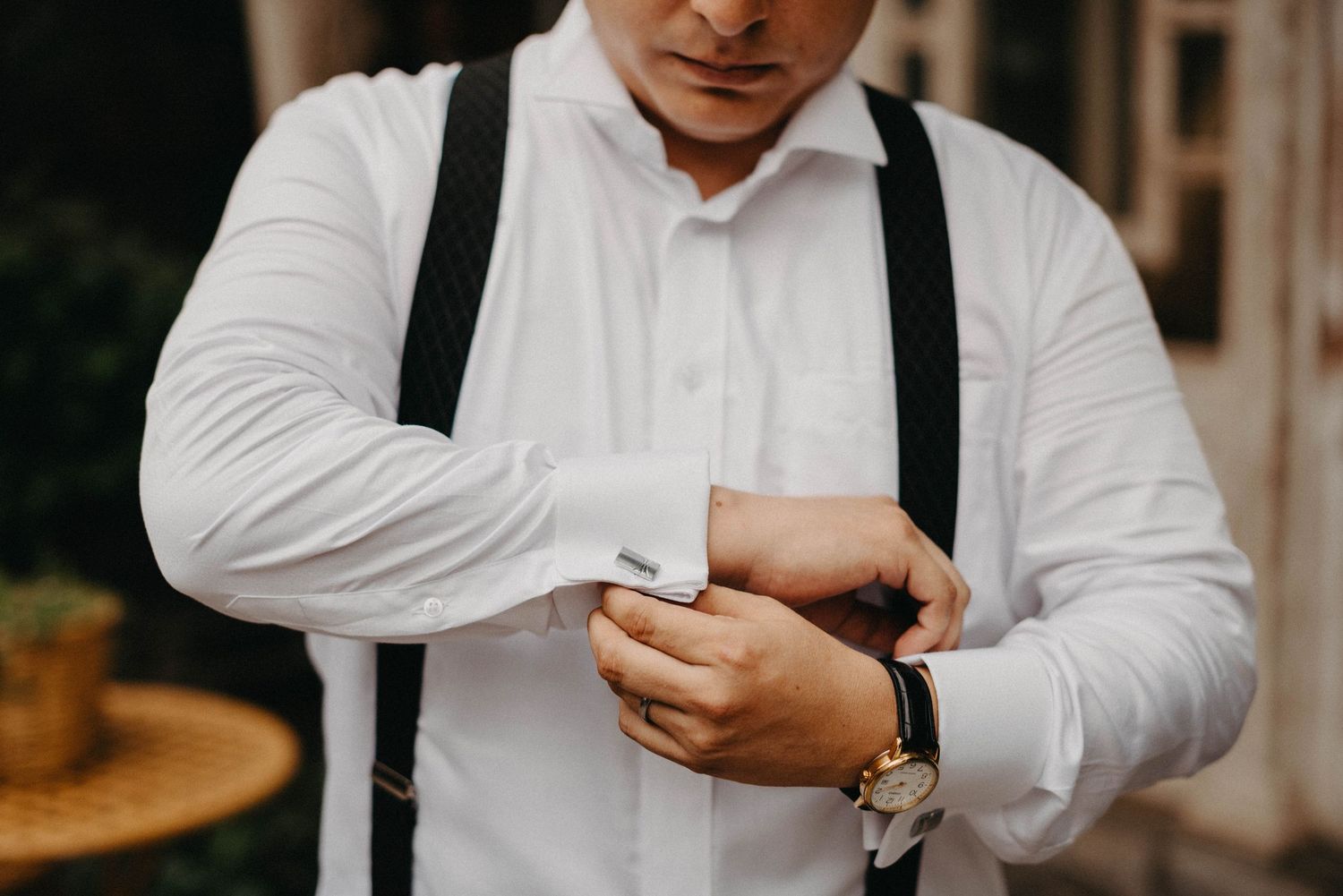 A person wearing a white dress shirt and black suspenders adjusts their cuffs in a formal setting.