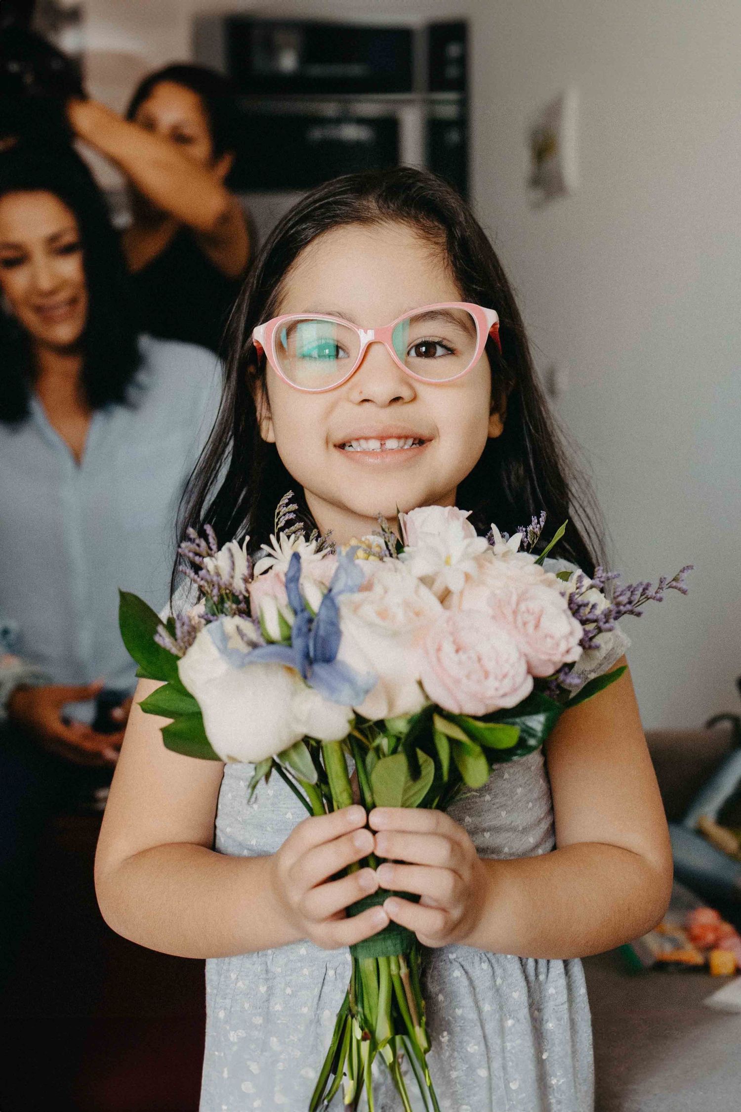 Young flower girl smiles while holding pastel bouquet during wedding preparations.