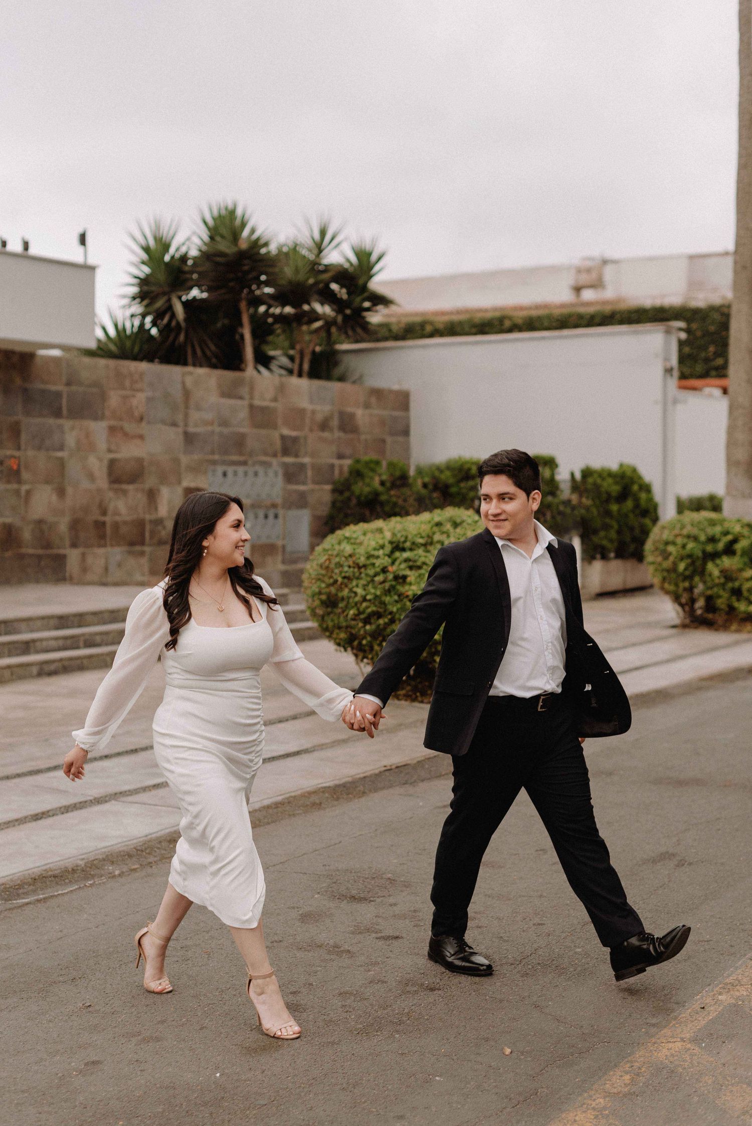 A playful couple holding hands while walking through an urban setting with stone walls.