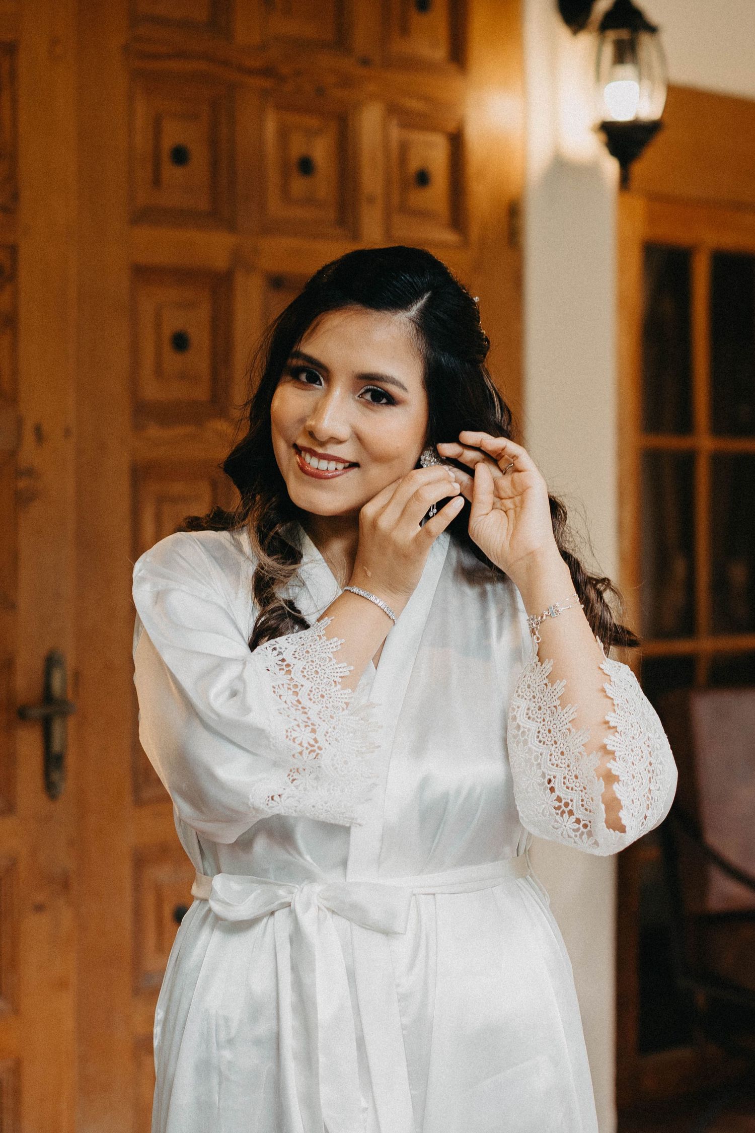 A person in a white lace-trimmed robe adjusts their earrings while getting ready in a warmly lit room.