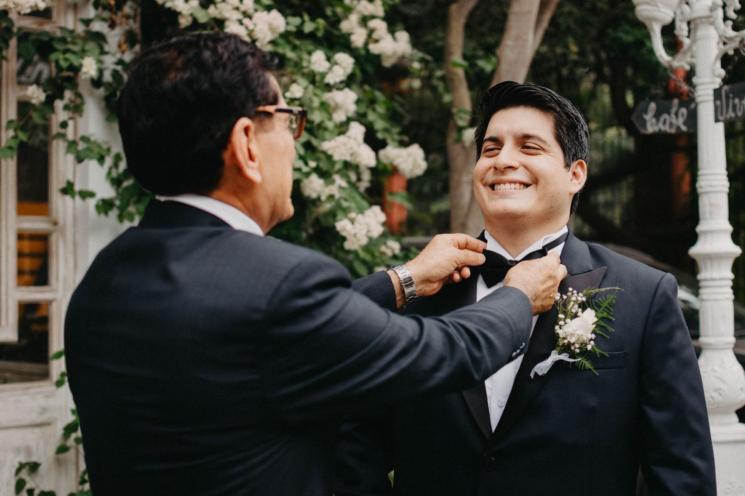 Someone helps adjust a bowtie on a groom wearing a black tuxedo during an outdoor wedding ceremony.