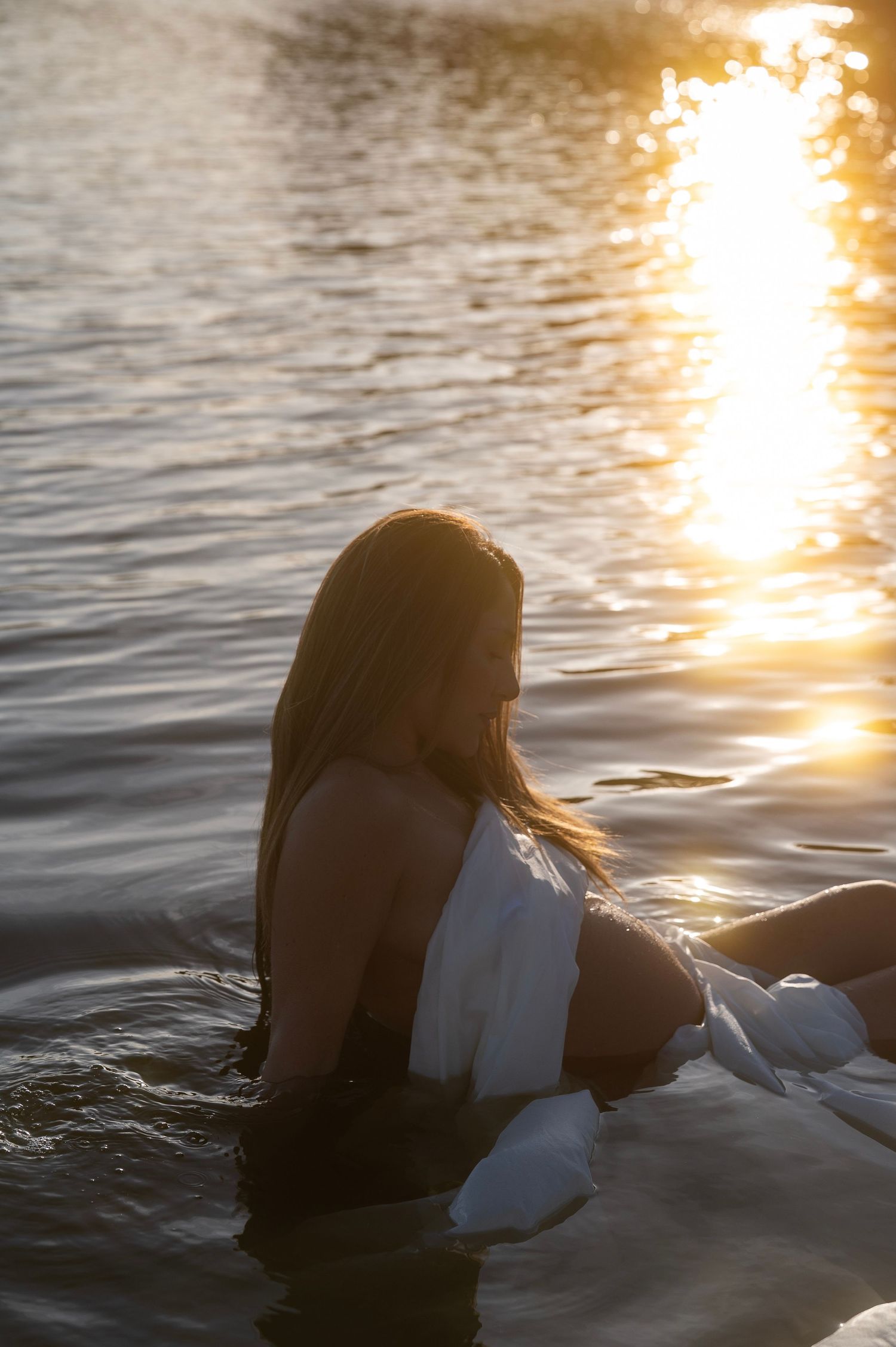 Pregnant woman sitting in golden sunlit water holding baby bump with white fabric draped around her.