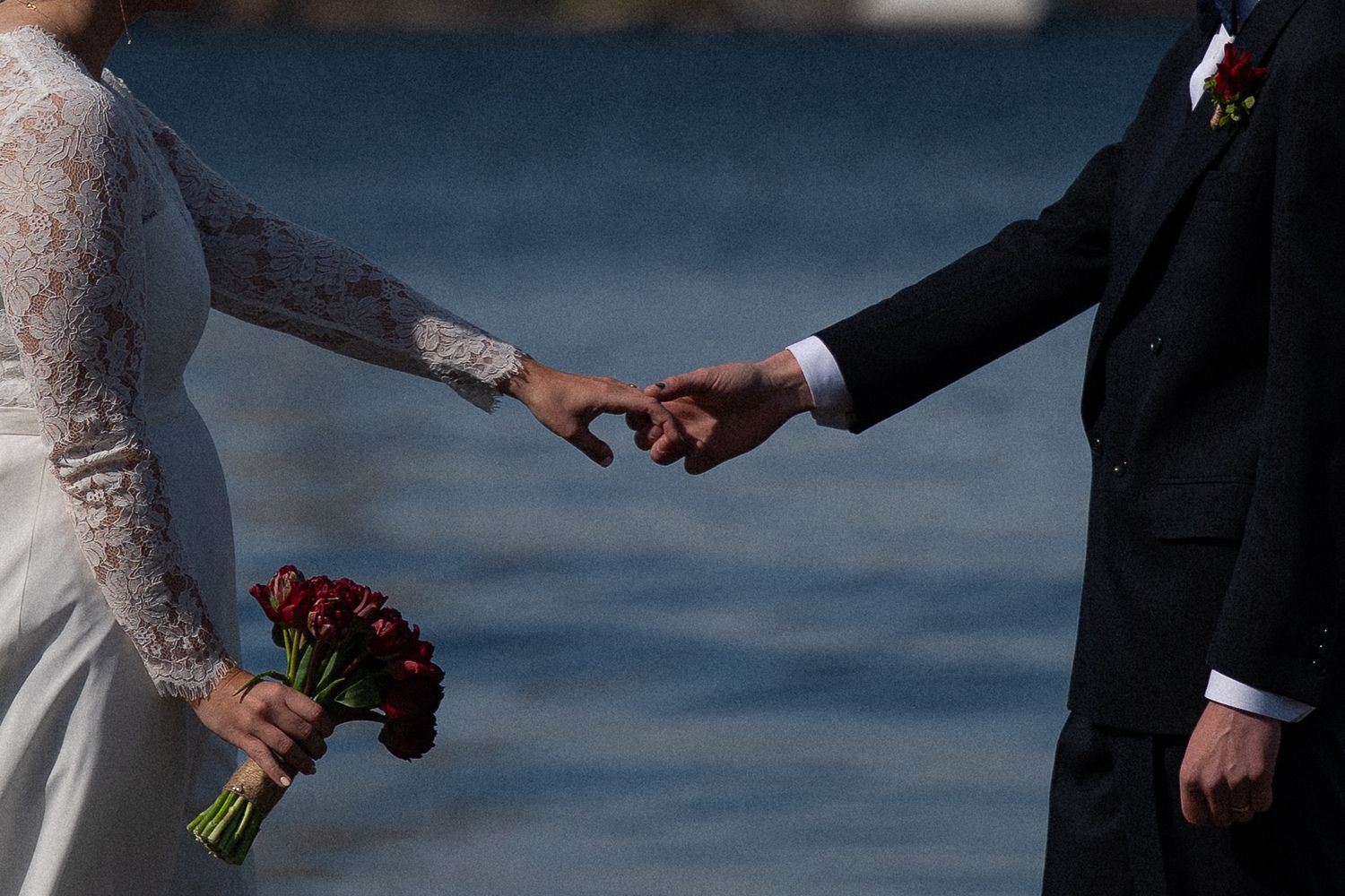 Bride and groom gently linking fingers outdoors near water, bride holding red rose bouquet in lace wedding dress.
