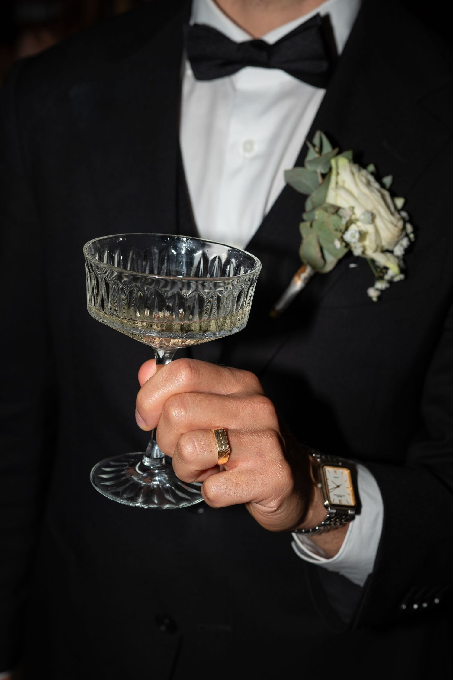 Groom in black tuxedo with boutonniere holding crystal coupe glass, wearing gold wedding ring and watch.