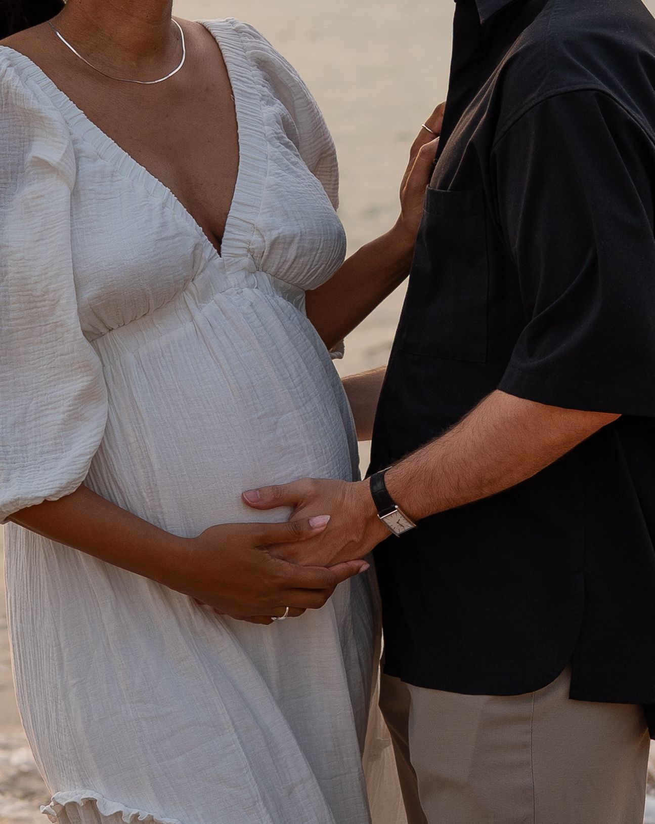 Pregnant woman in white dress with partner in black shirt gently touching baby bump outdoors.
