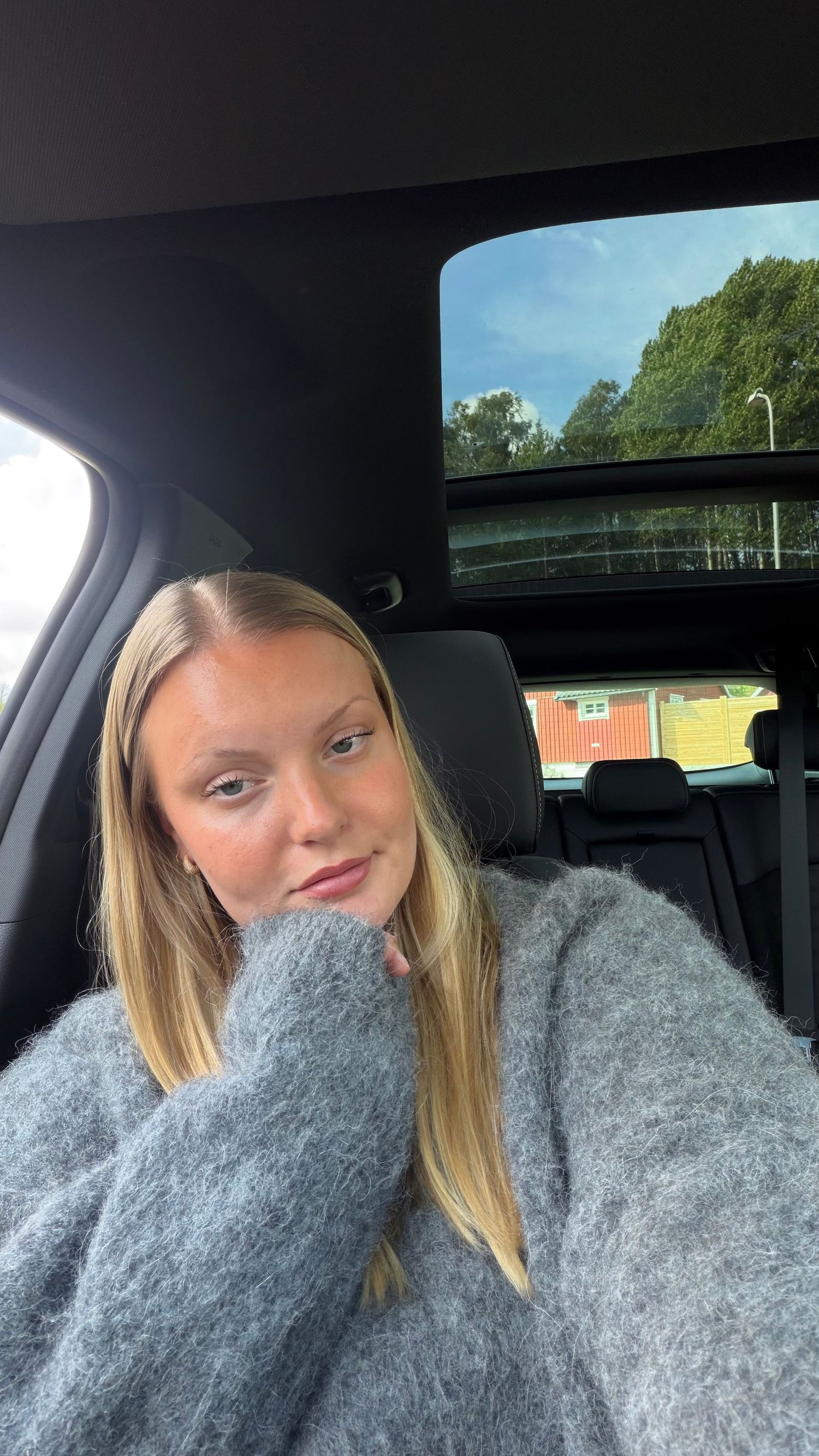 Young blonde woman in gray fuzzy sweater taking a selfie in a car with sunroof and trees visible outside.