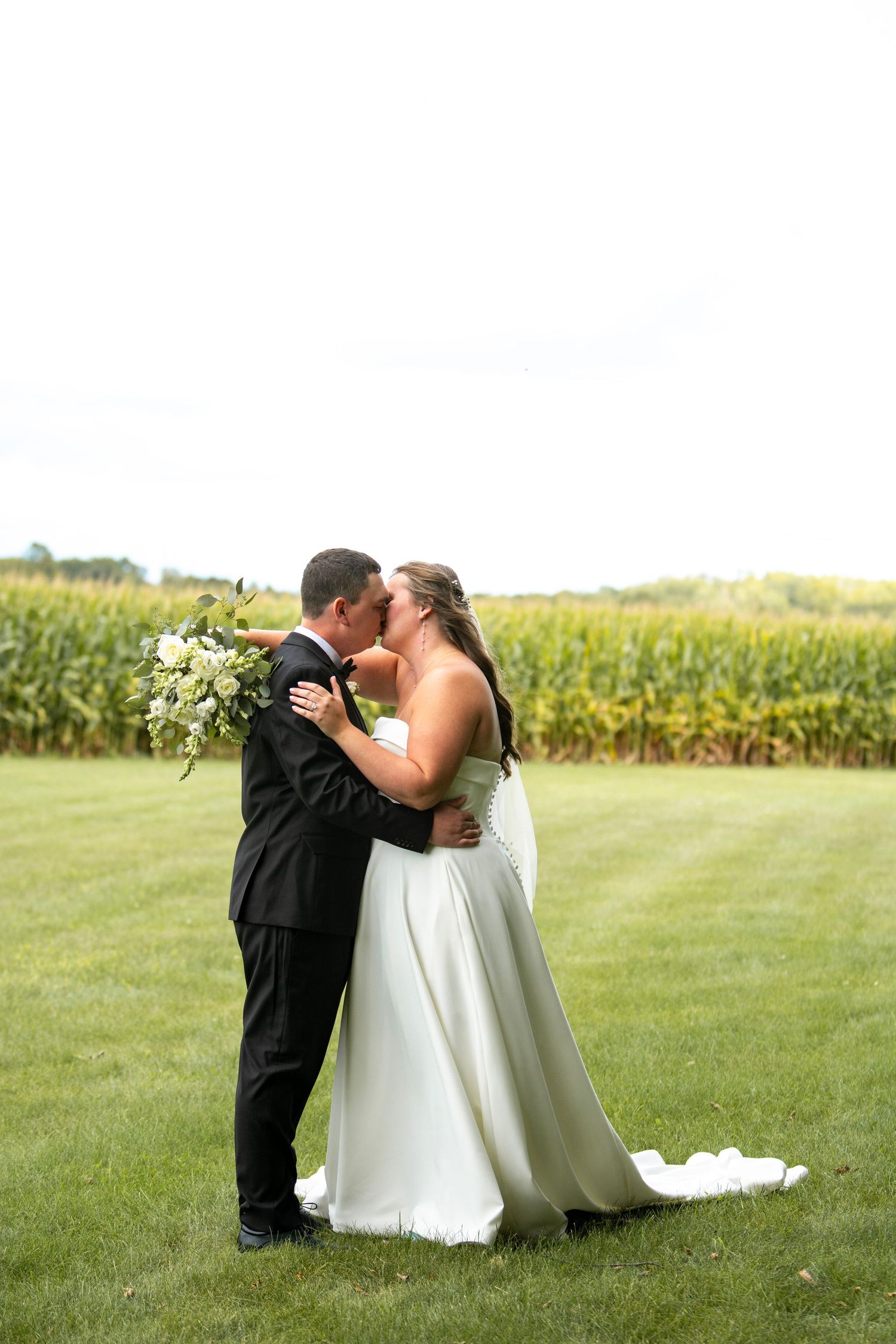 Newlywed couple shares intimate moment sitting together in grassy field with cornfield backdrop.