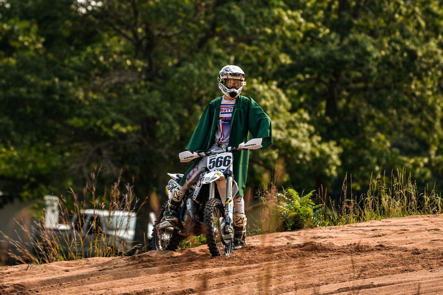 Dirt bike rider performing stunts on a dusty off-road track surrounded by trees.