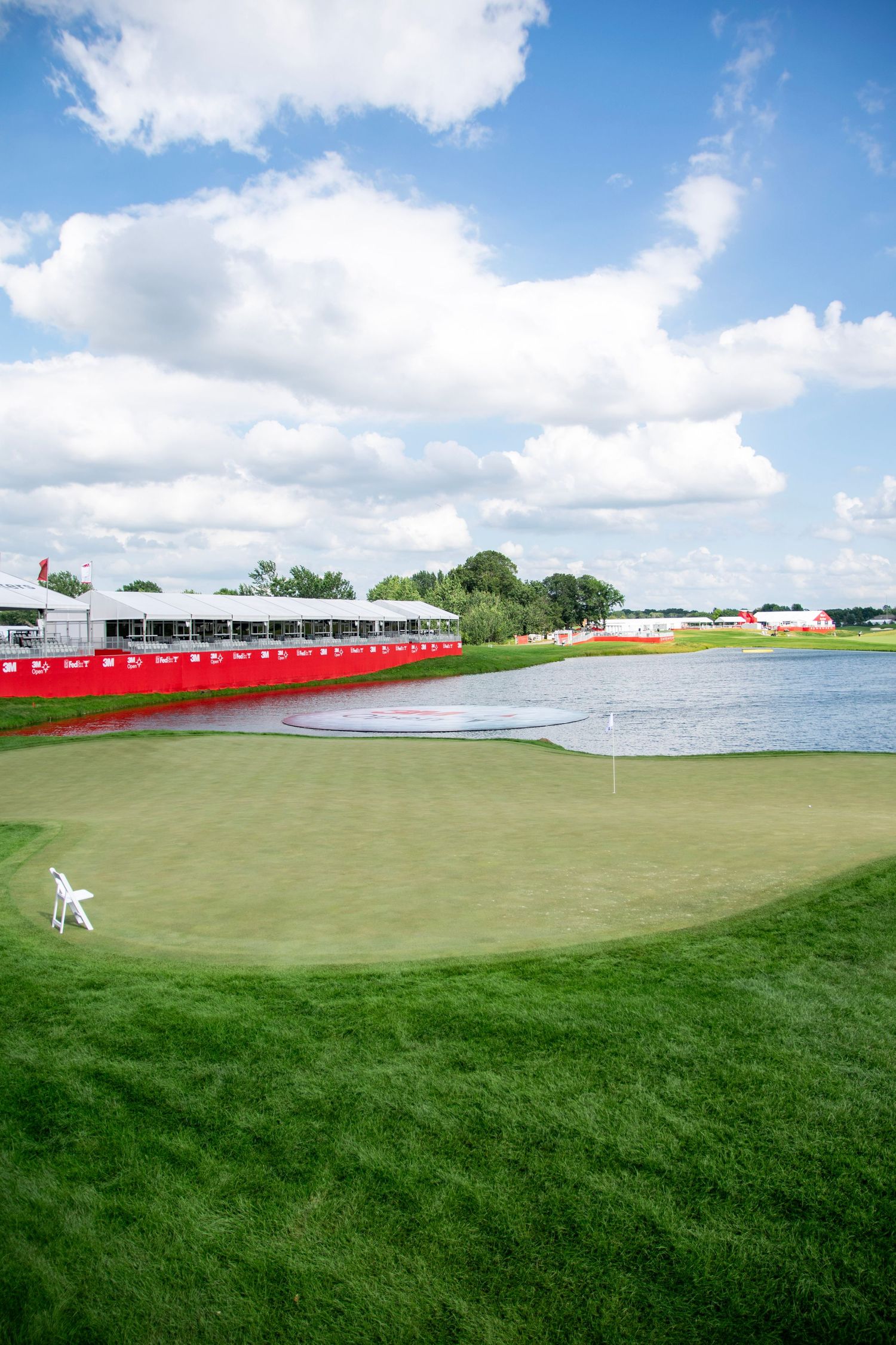 A panoramic view of a pristine golf course with lush green fairways and white tents under blue skies with puffy clouds.