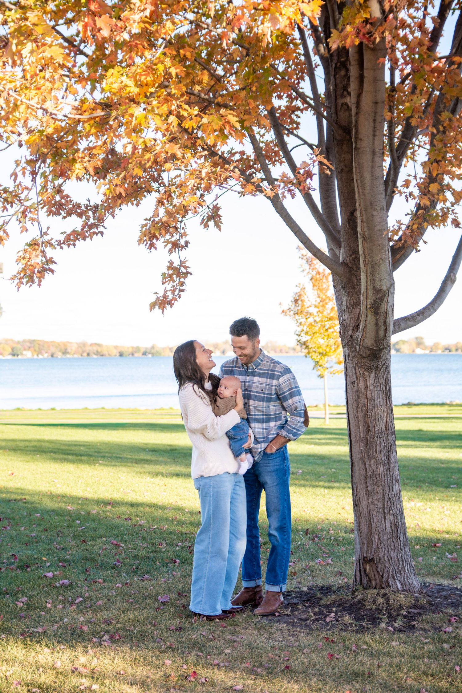 Couple poses together under fall foliage by lakeside on sunny autumn day.