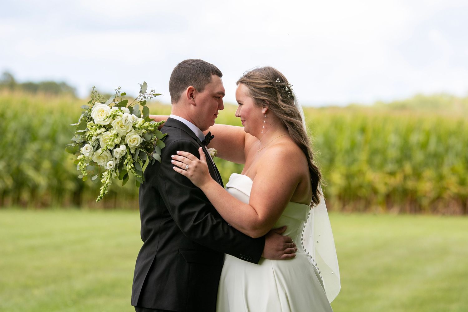 Bride and groom share a romantic moment in front of a lush cornfield, bride holding a white floral bouquet.