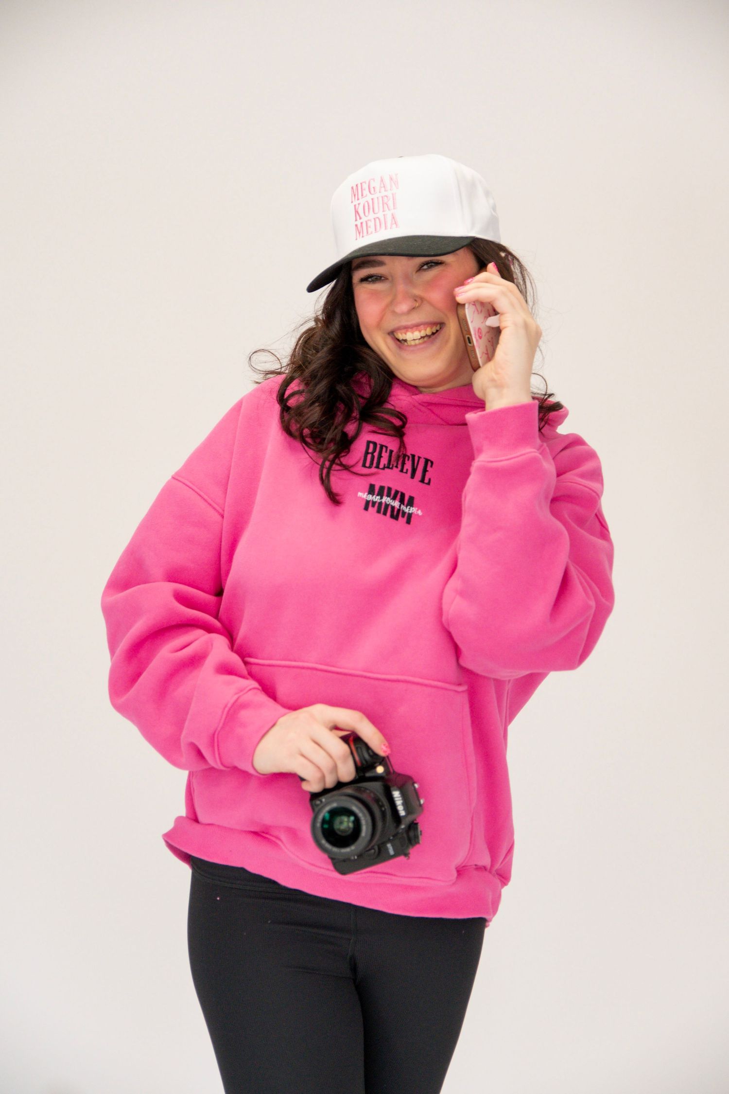 Person in bright pink sweatshirt and black pants holds camera while wearing white baseball cap against light background.