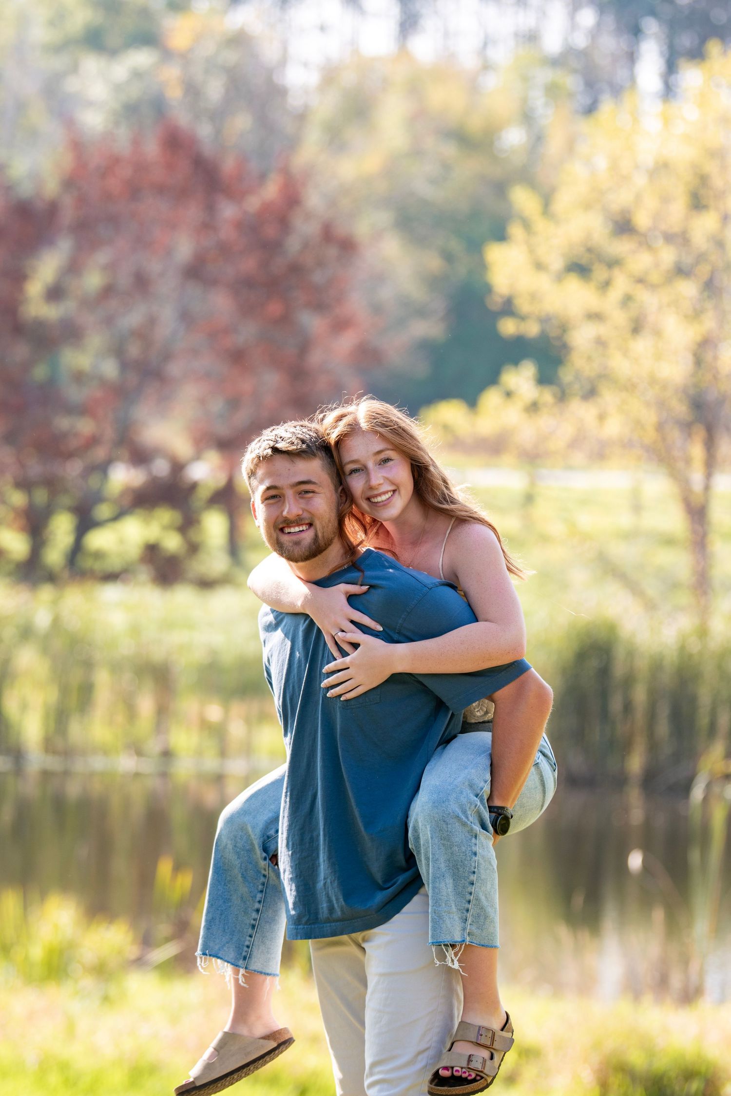 Playful couple enjoys piggyback ride near lake during autumn sunset.