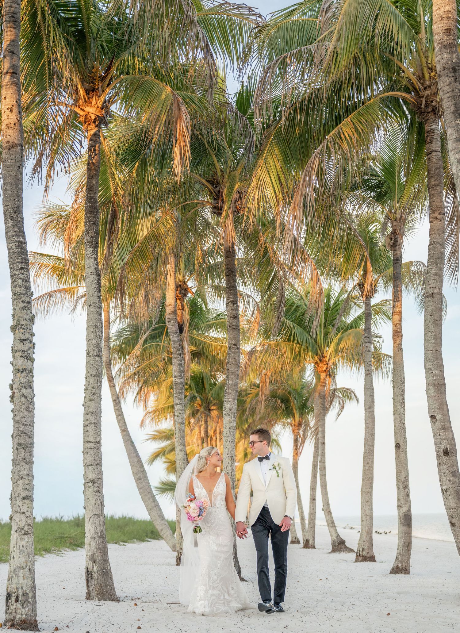 Bride and groom walk hand-in-hand on a sandy beach surrounded by golden palm trees at sunset.