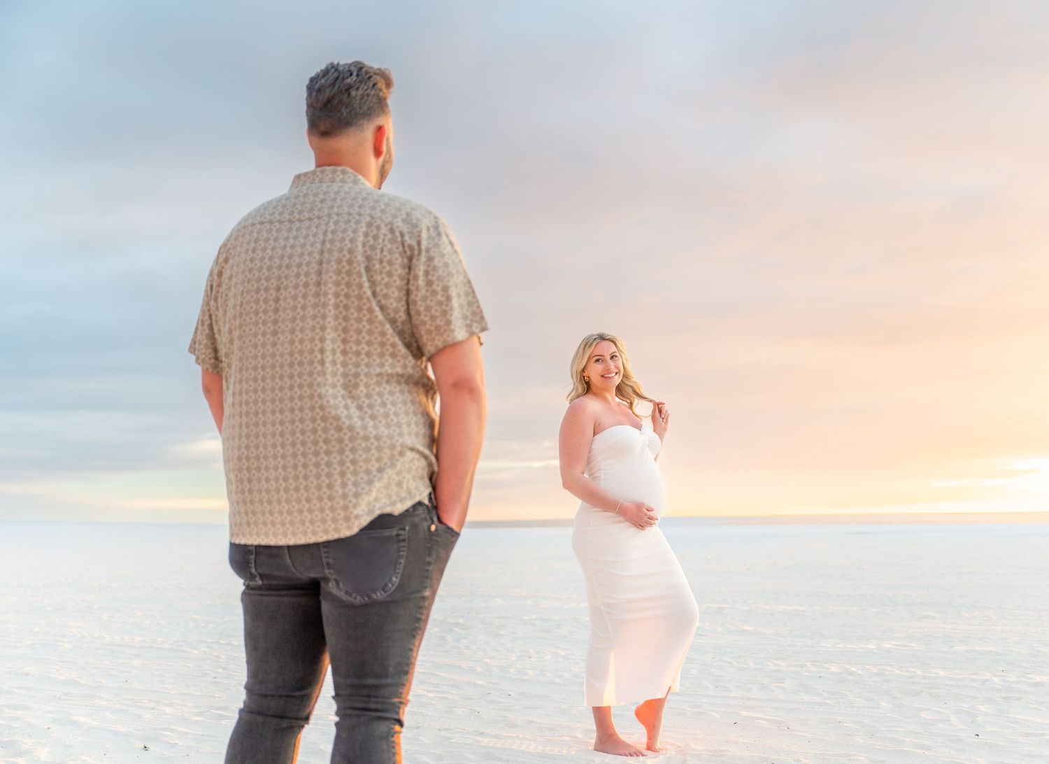 Couple poses on white salt flats at sunset, pregnant woman in white dress cradles baby bump while partner stands nearby.