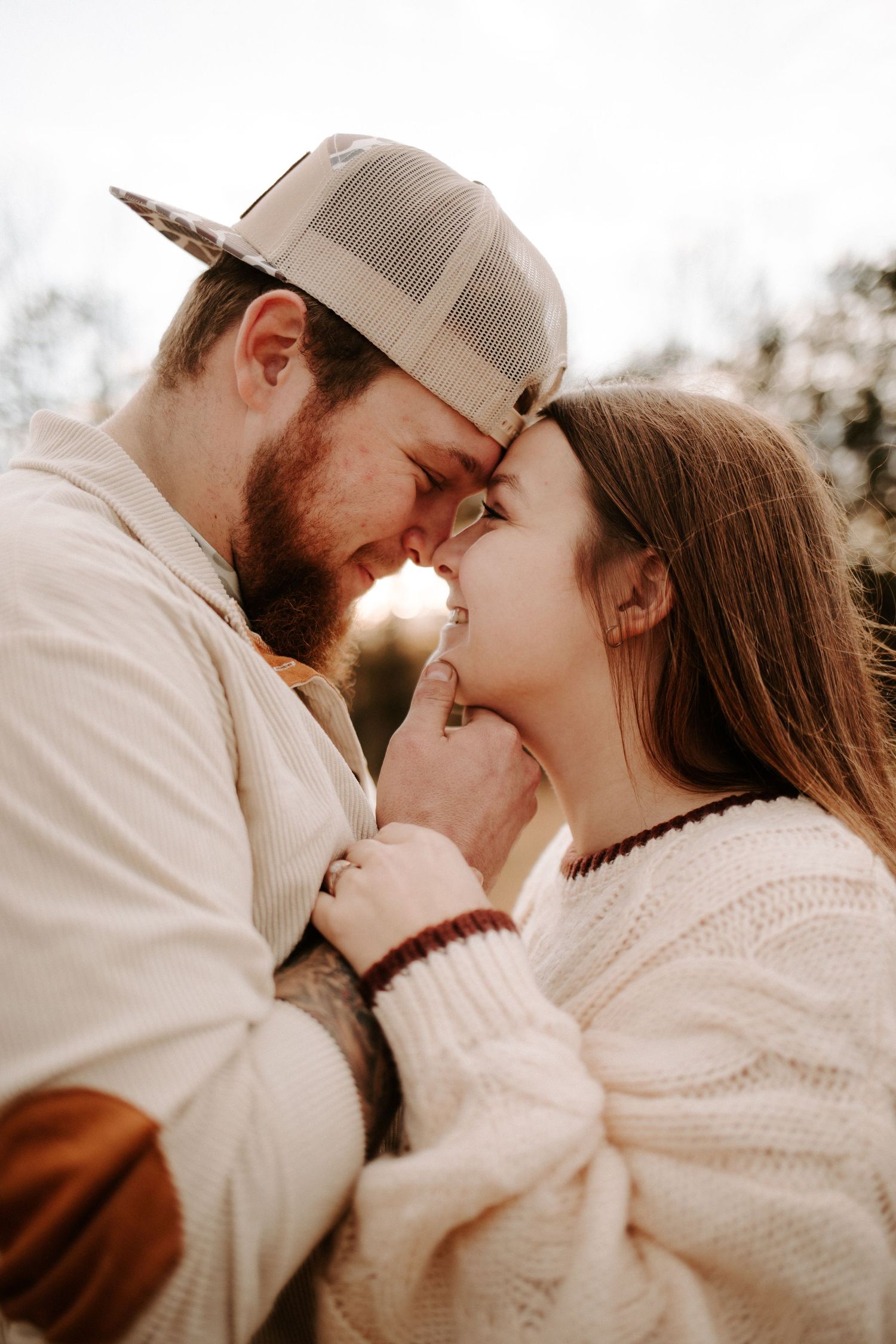 A bearded man in a cap and tattooed woman in a white sweater share a romantic forehead touch outdoors.