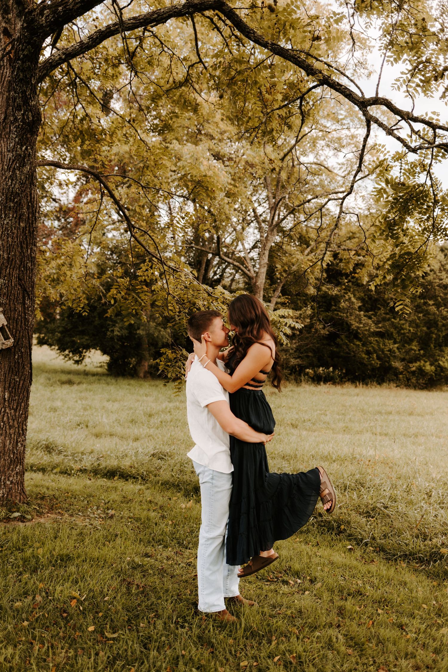 A couple shares a romantic embrace under a large tree in a lush green field, the man lifting the woman in a black dress.