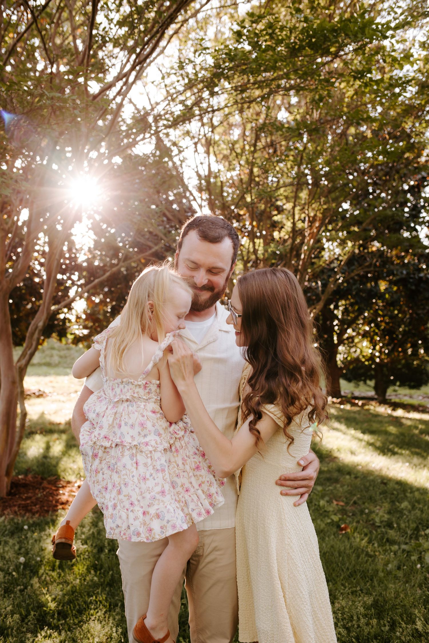 A family of three shares a tender moment outdoors, with golden sunlight filtering through lush green trees.