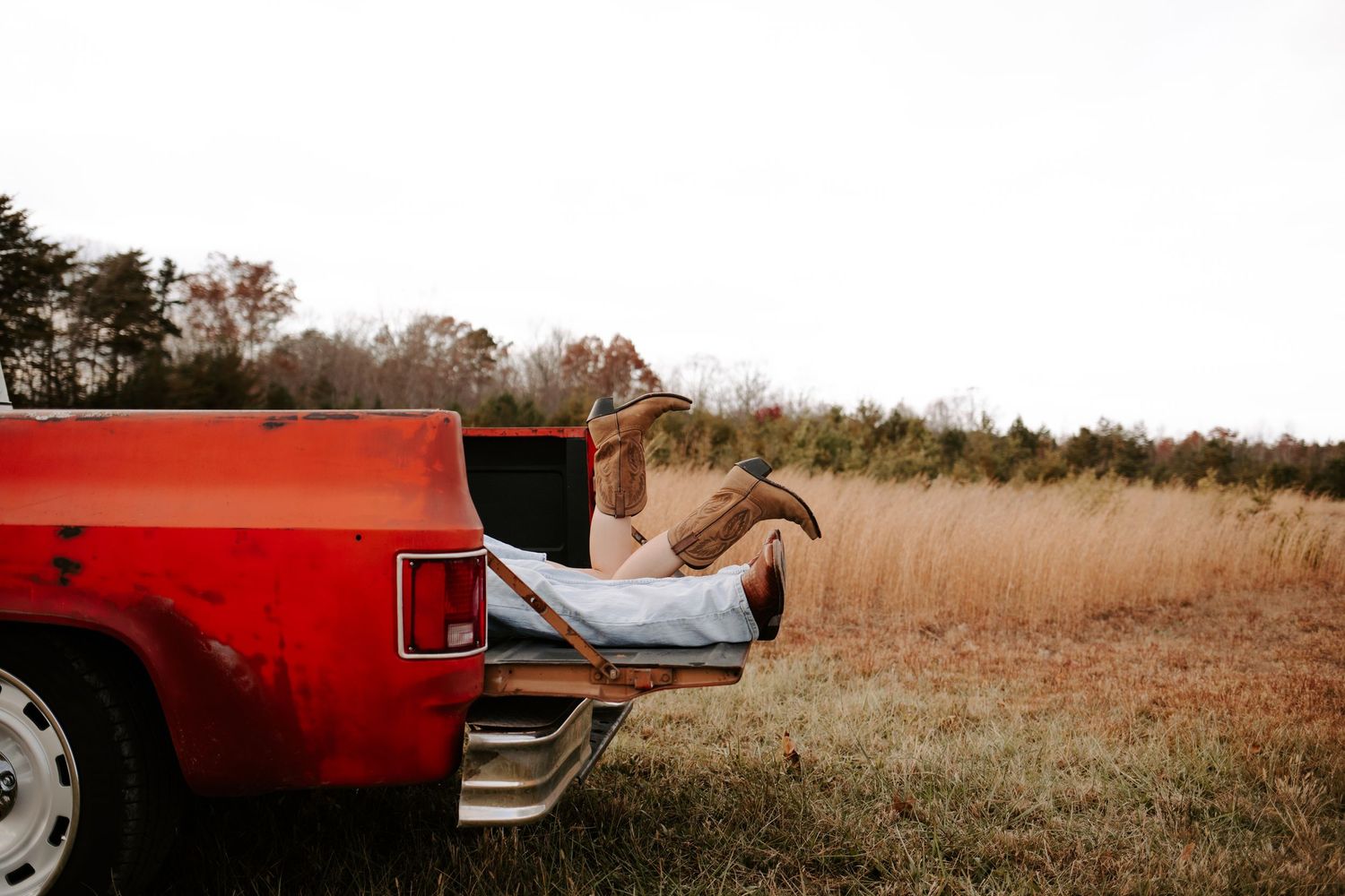 Person relaxing in the bed of a red pickup truck in a rural field, wearing cowboy boots, with golden grass in the background.